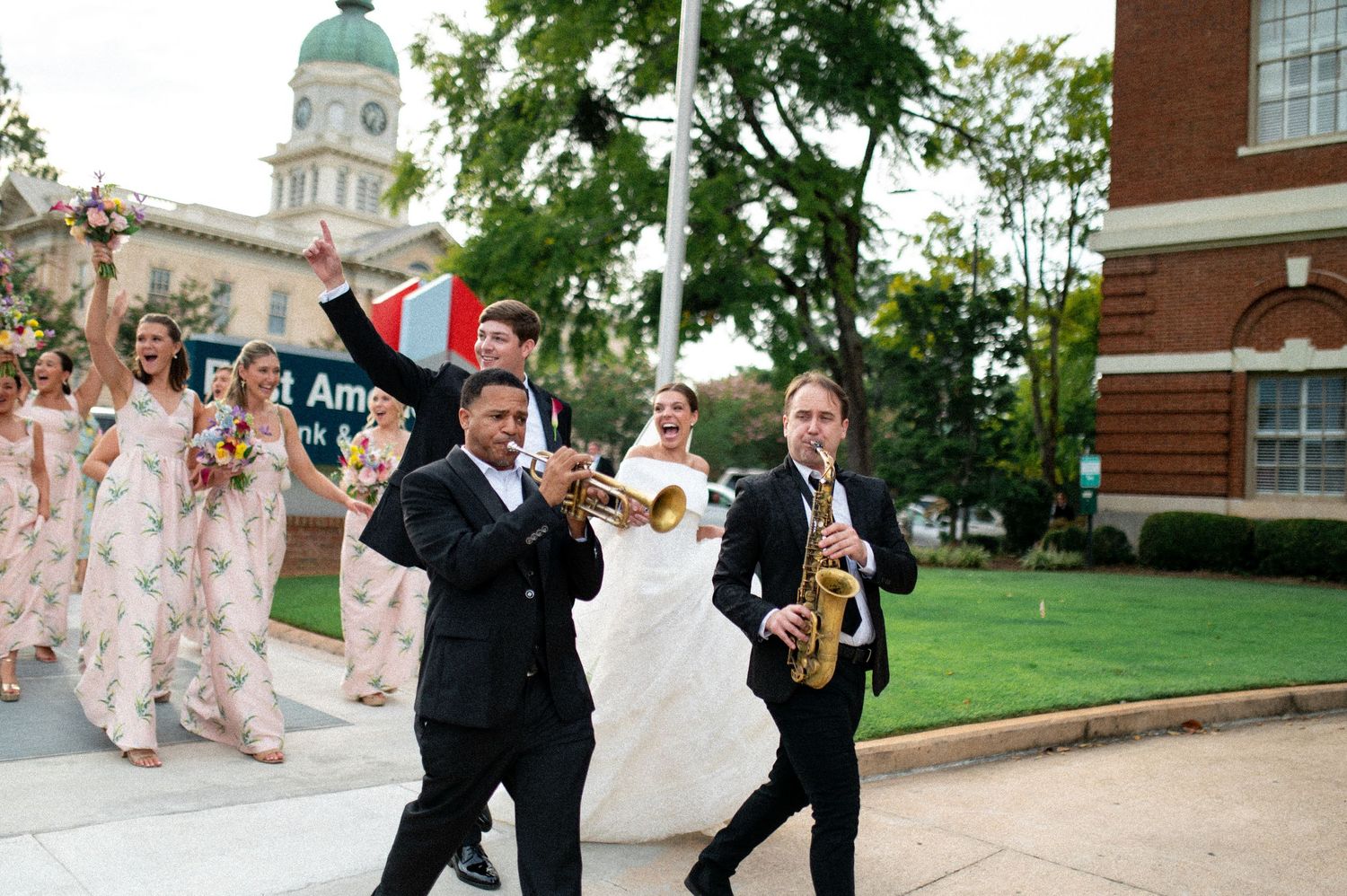 Bride leading a festive second line parade with brass band musicians through a city street.