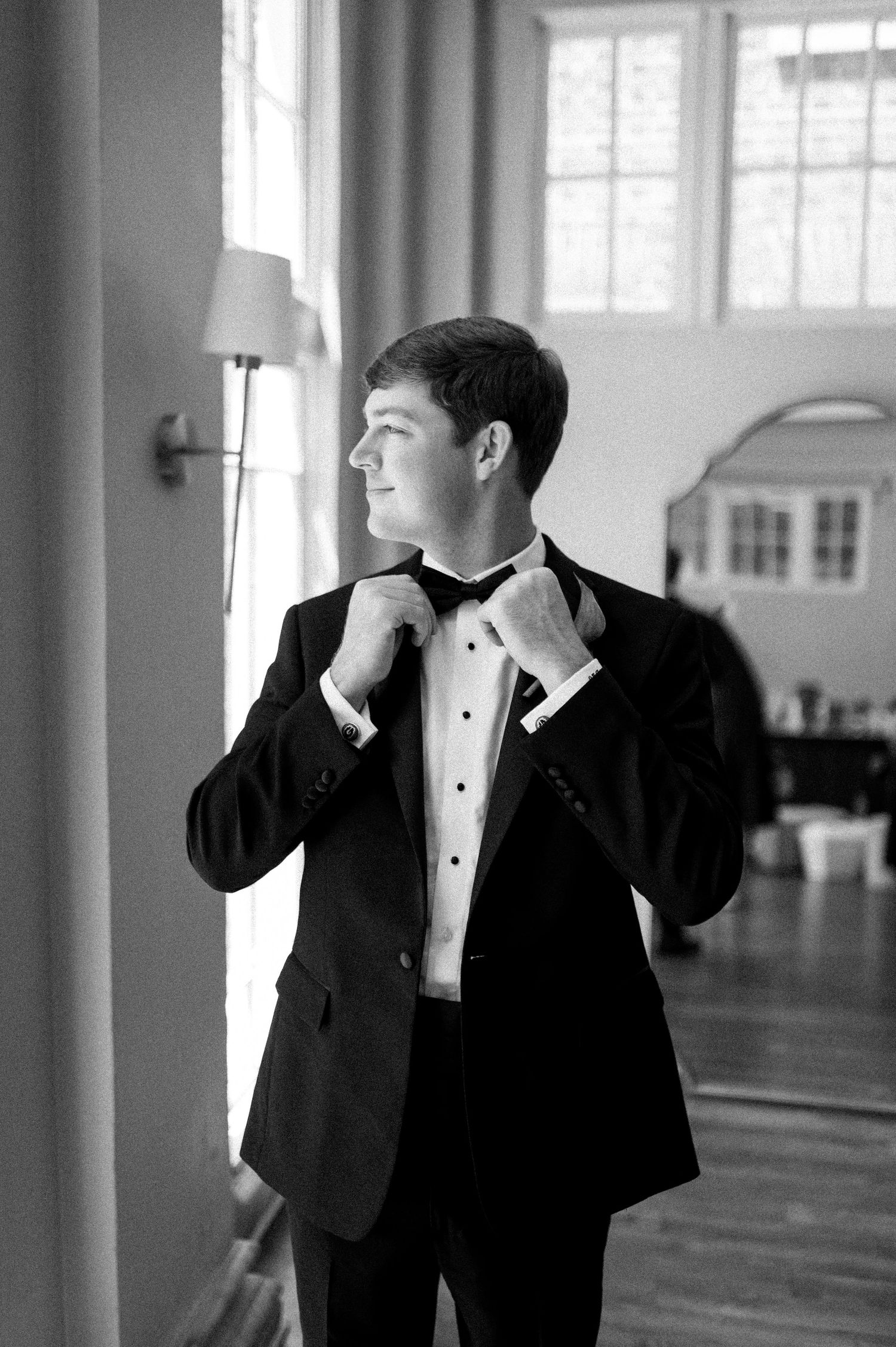 Groom in black tuxedo adjusting bow tie near window in black and white wedding photo.