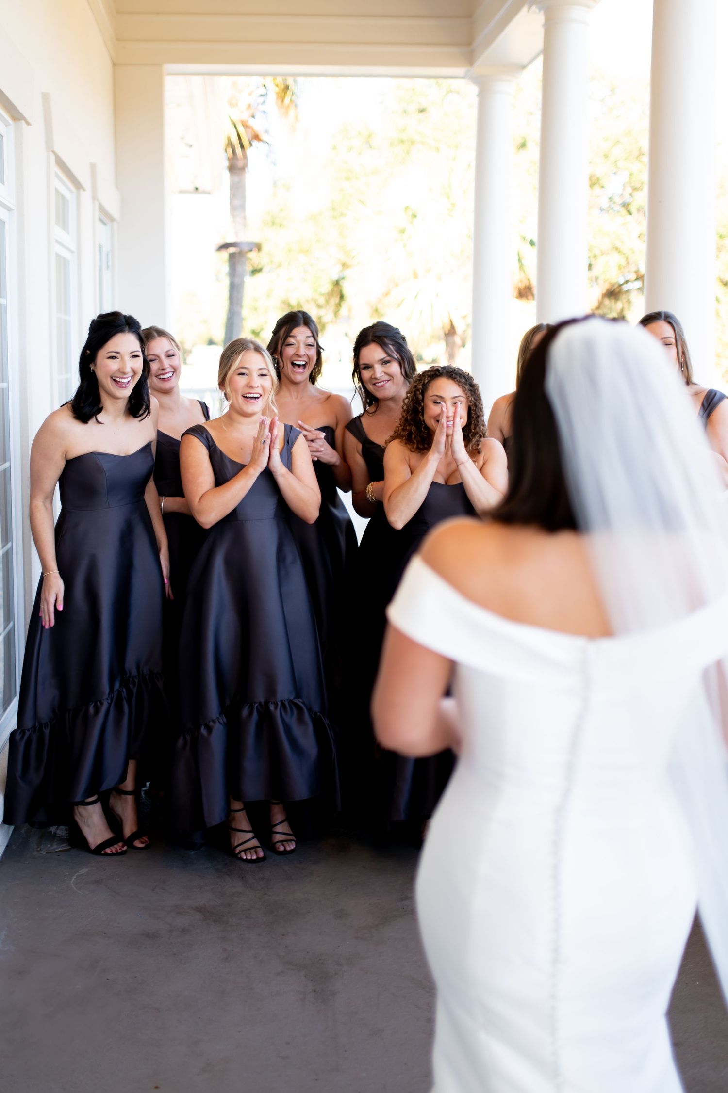 Wedding party in black dresses celebrating with bride in white on covered porch.