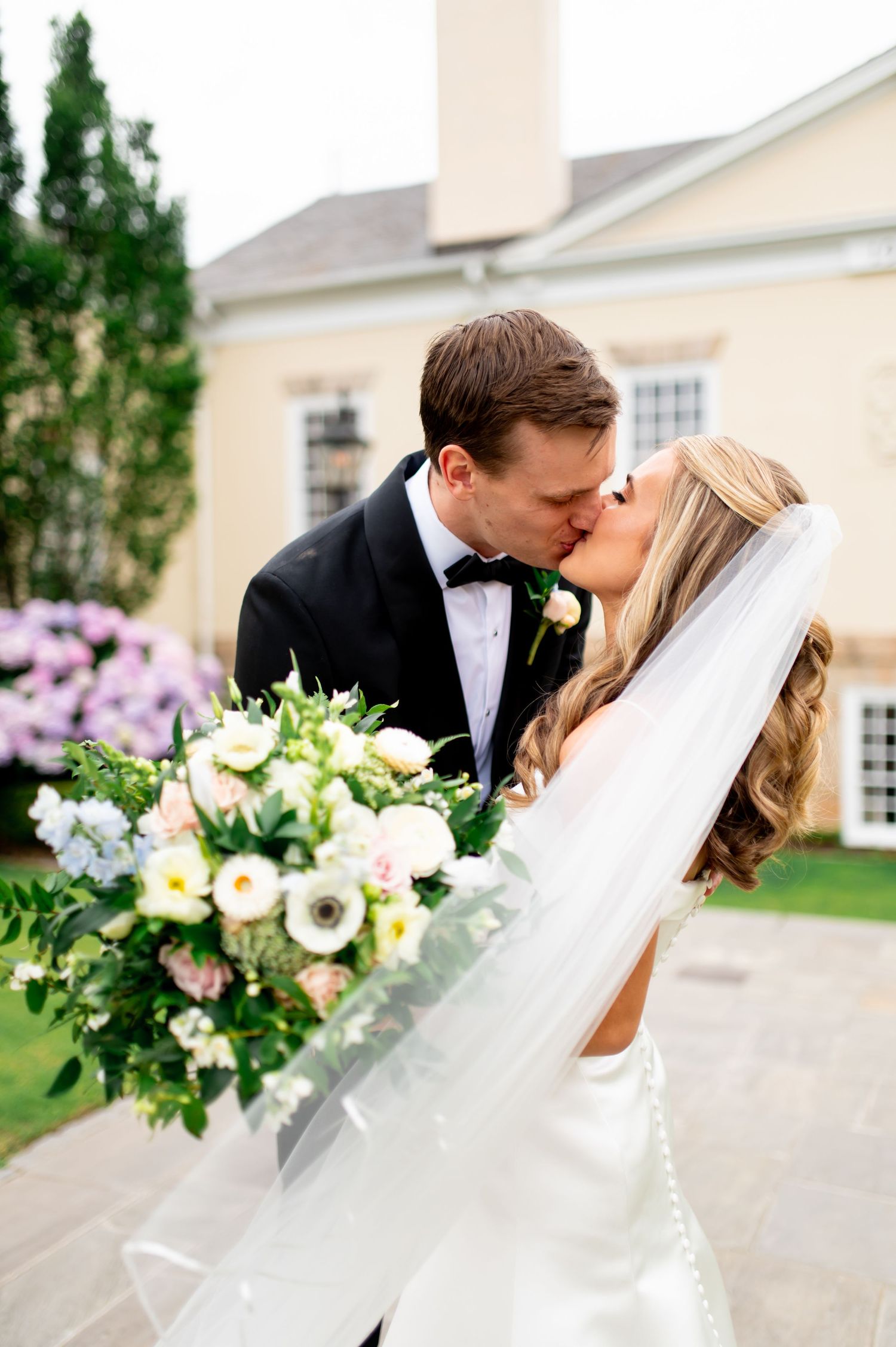Romantic wedding kiss captured in front of elegant building with floral bouquet.