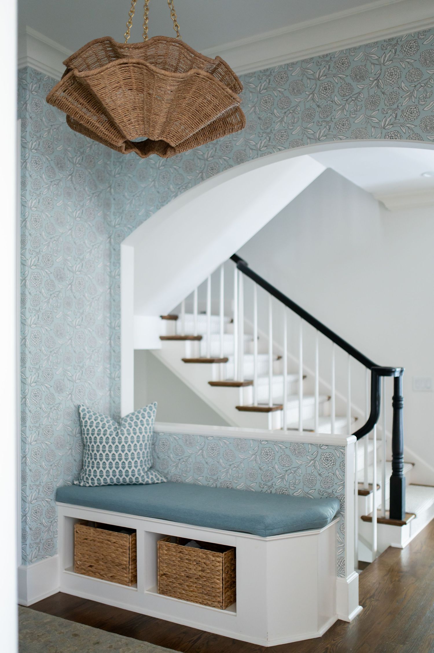 Coastal entryway with blue floral wallpaper, built-in bench with wicker baskets, rattan chandelier, and white staircase.