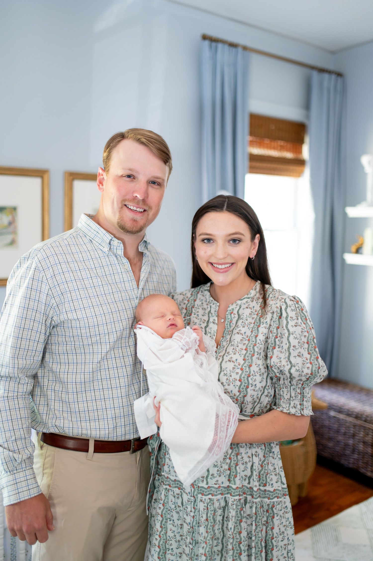 A couple stands in a bright living room holding their newborn baby dressed in white.