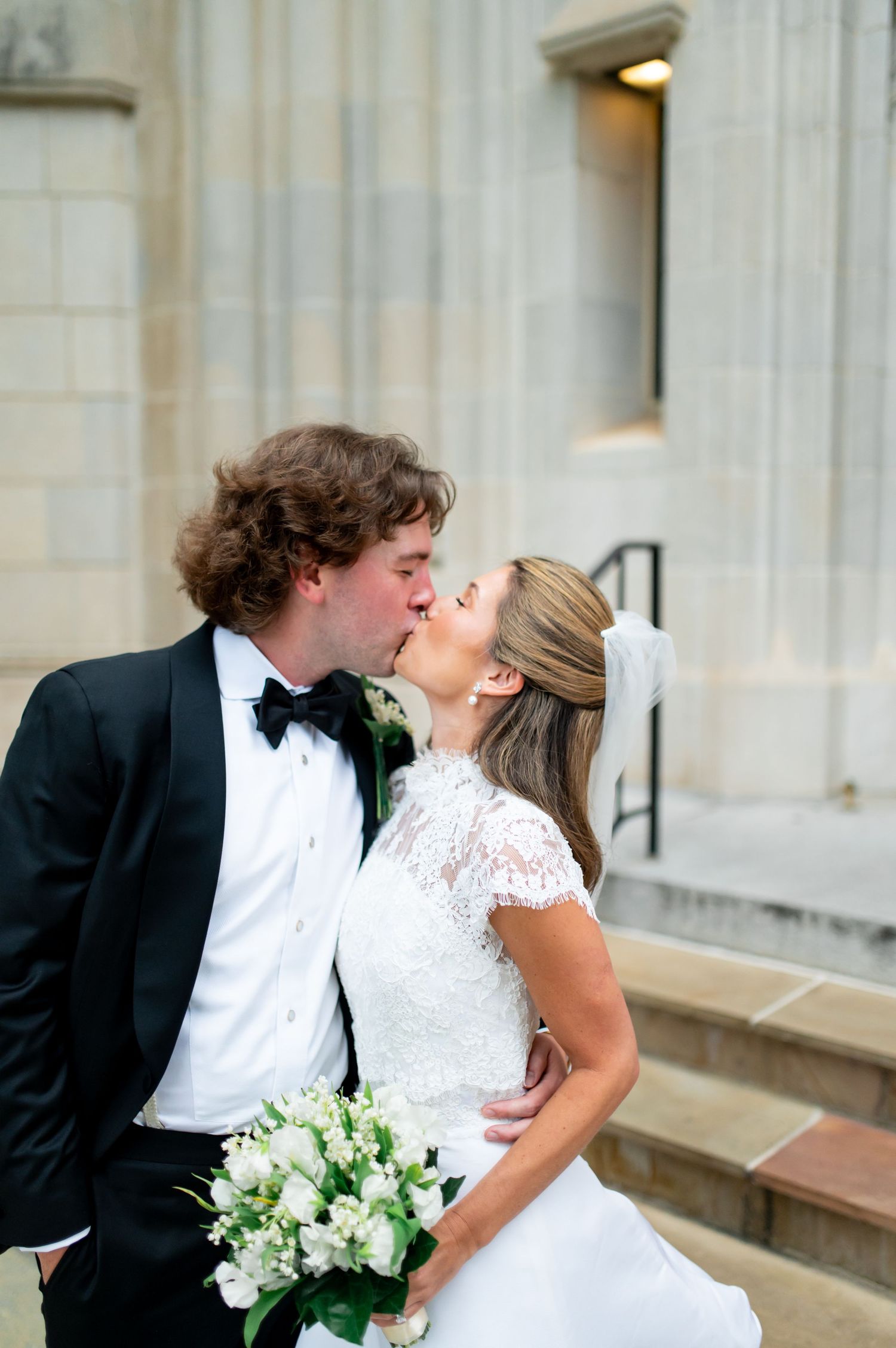 A romantic wedding kiss captured in front of a stone church entrance with white bouquet.