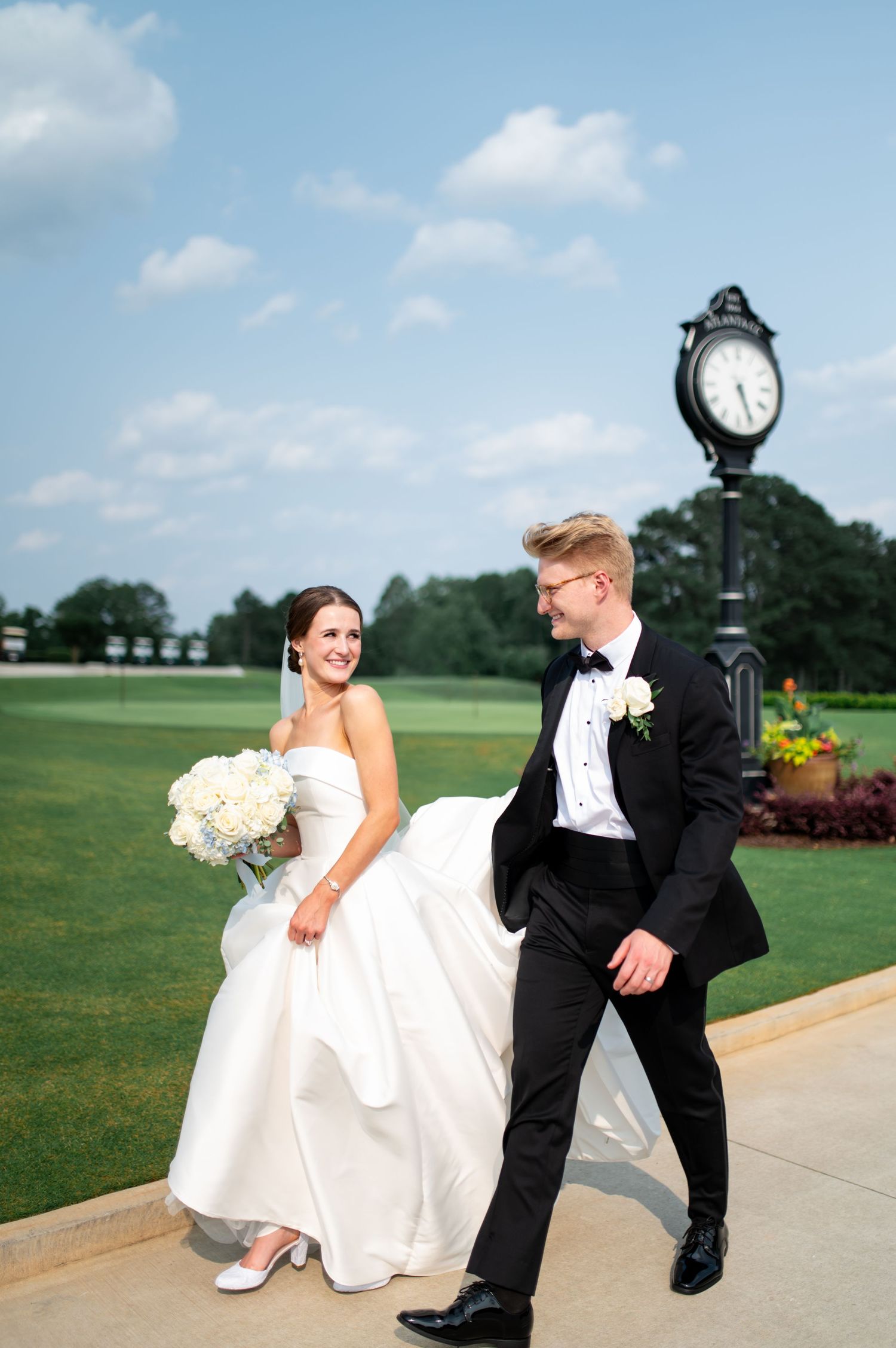 Playful wedding portraits on a sunny day with outdoor clock and landscaped grounds.