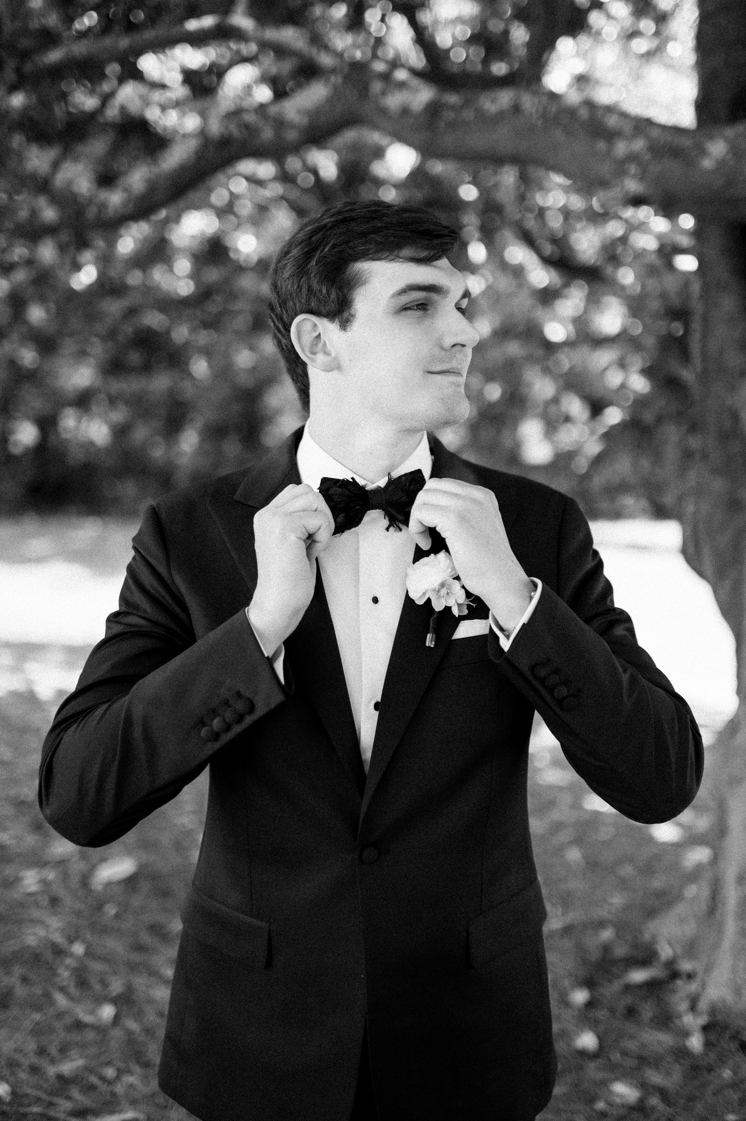 Dapper groom in black tuxedo adjusts bow tie outdoors in classic black and white portrait.