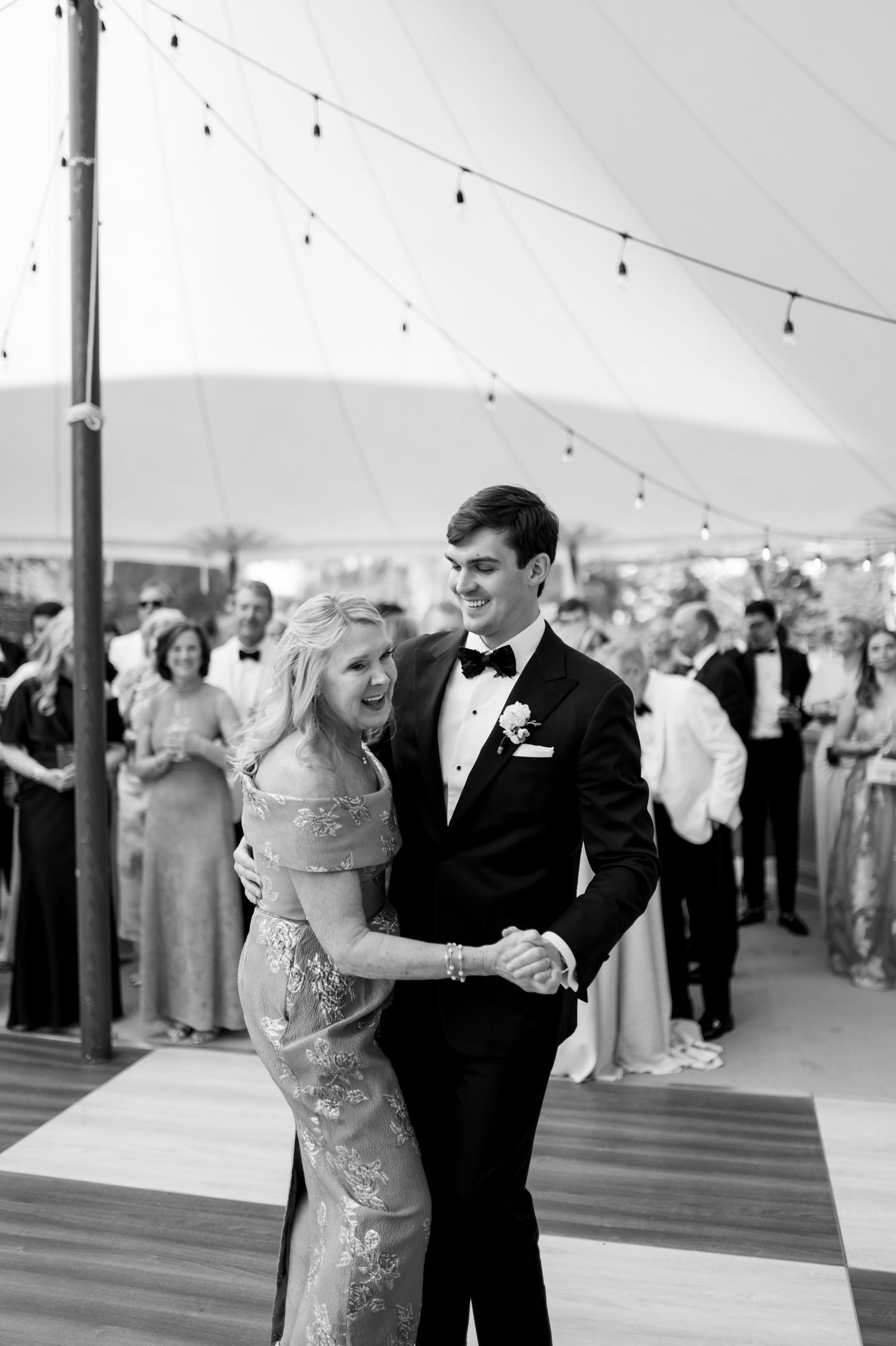 Black and white photo of groom in tuxedo sharing mother-son dance under tented reception venue.