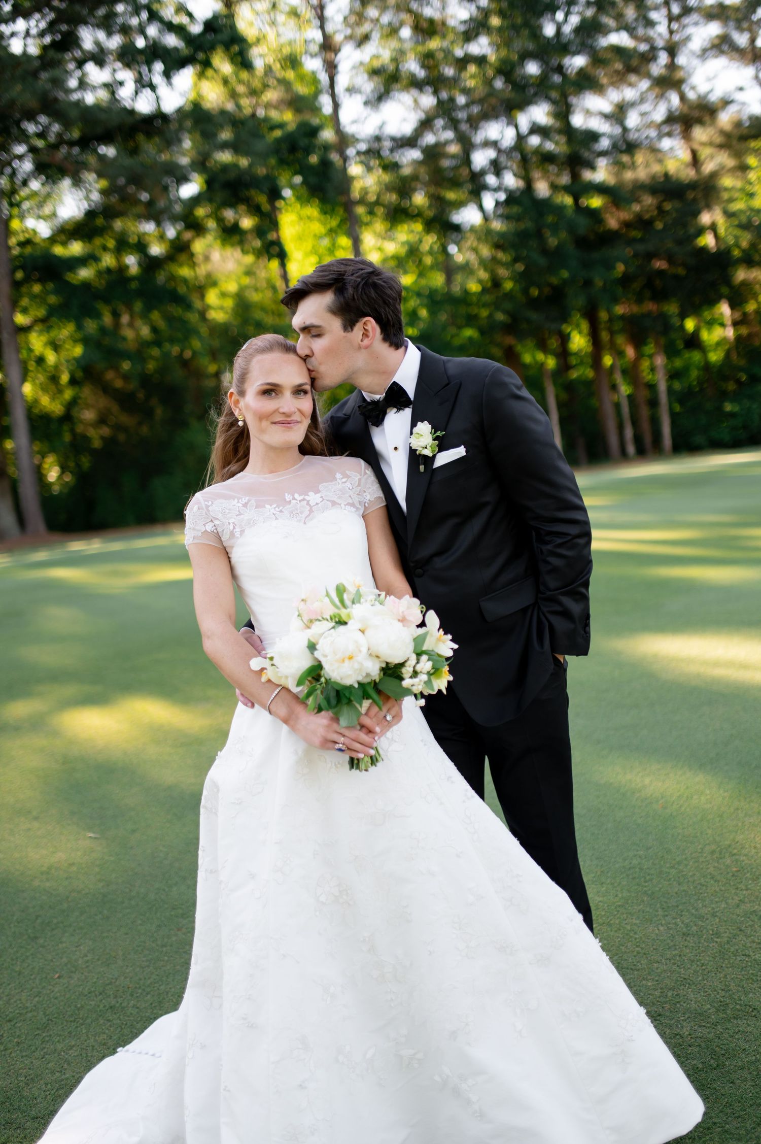 Groom kissing bride on cheek on golf course, bride holding white peony bouquet at sunset.