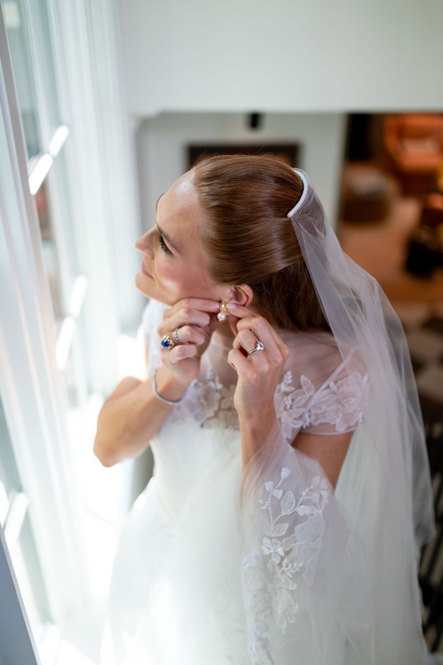 Bride in lace wedding dress and veil putting on earrings by a bright window on her wedding day.