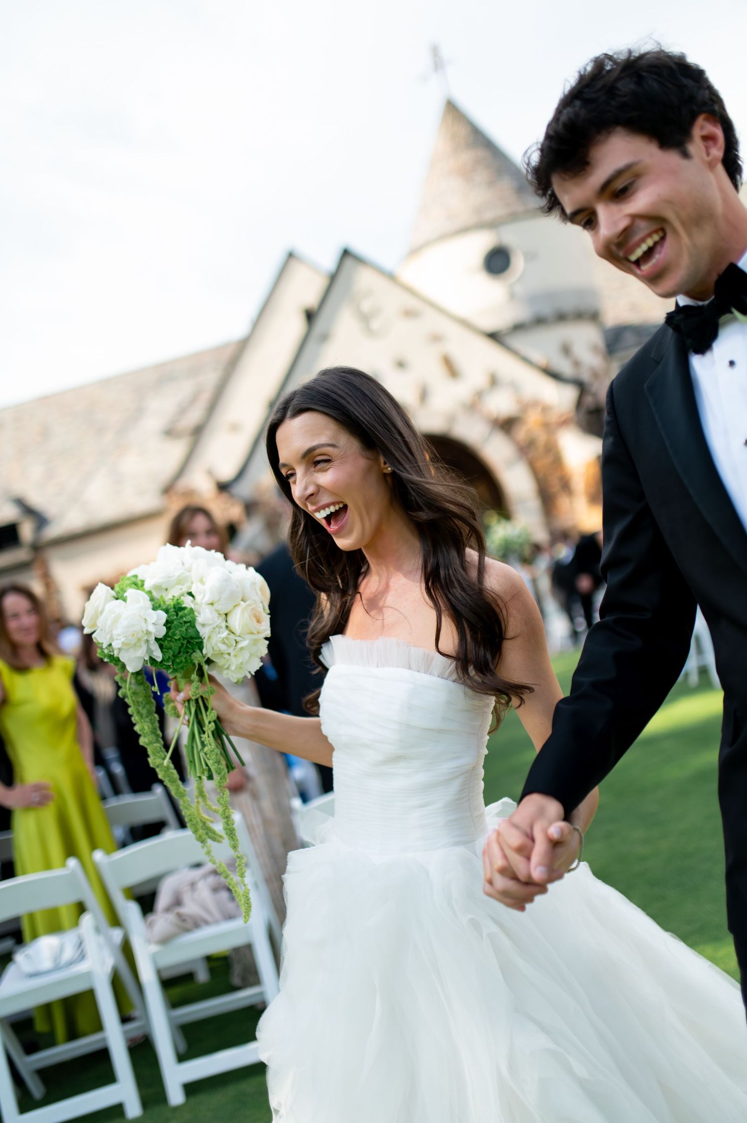 Joyful bride in white strapless gown holding bouquet walks with groom in tuxedo after outdoor church wedding ceremony.