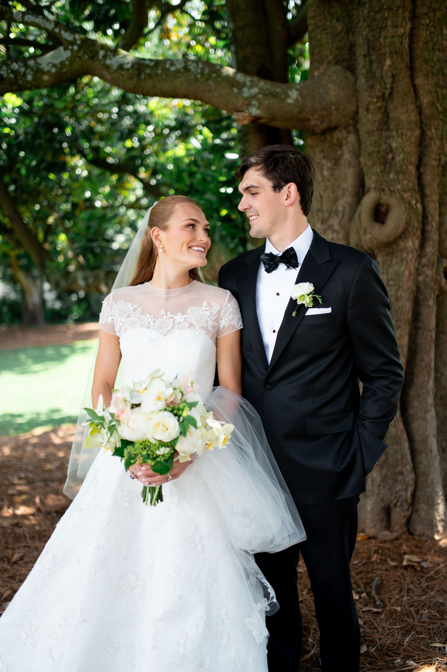 Smiling bride and groom share romantic moment under oak tree, bride holding white floral bouquet.