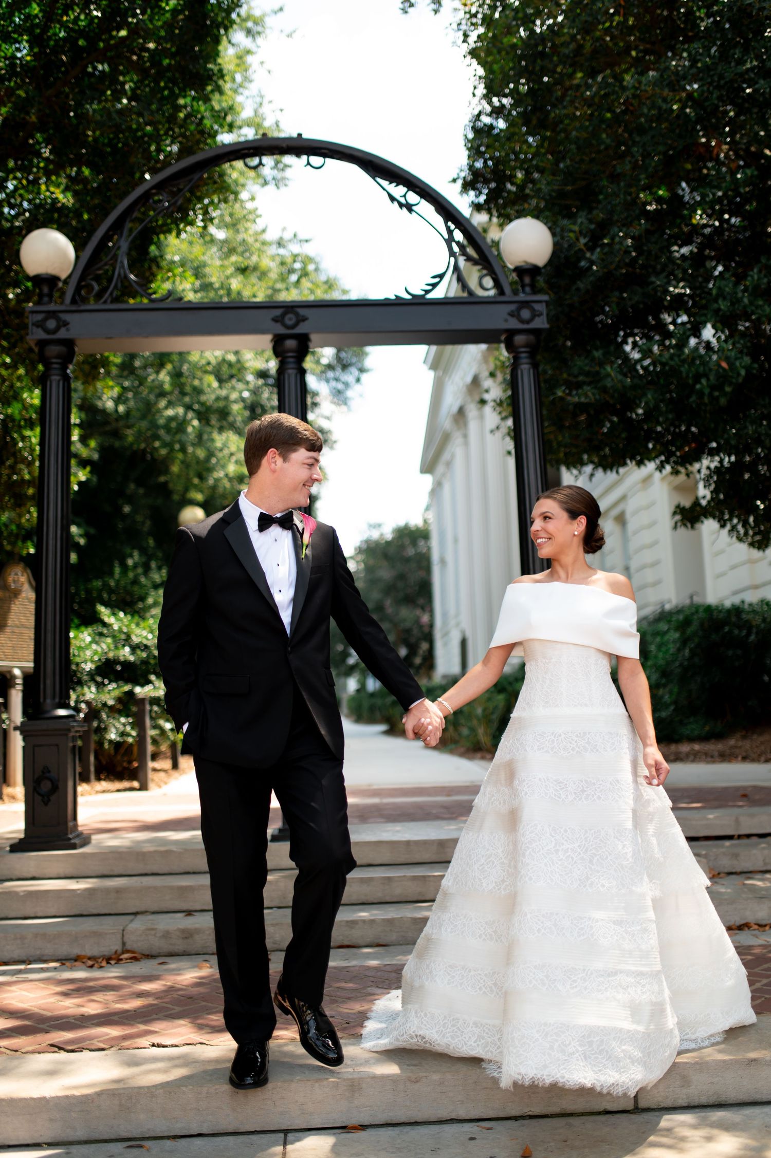 Bride in off-shoulder white gown and groom in black tuxedo hold hands walking under ornate arch outdoors.