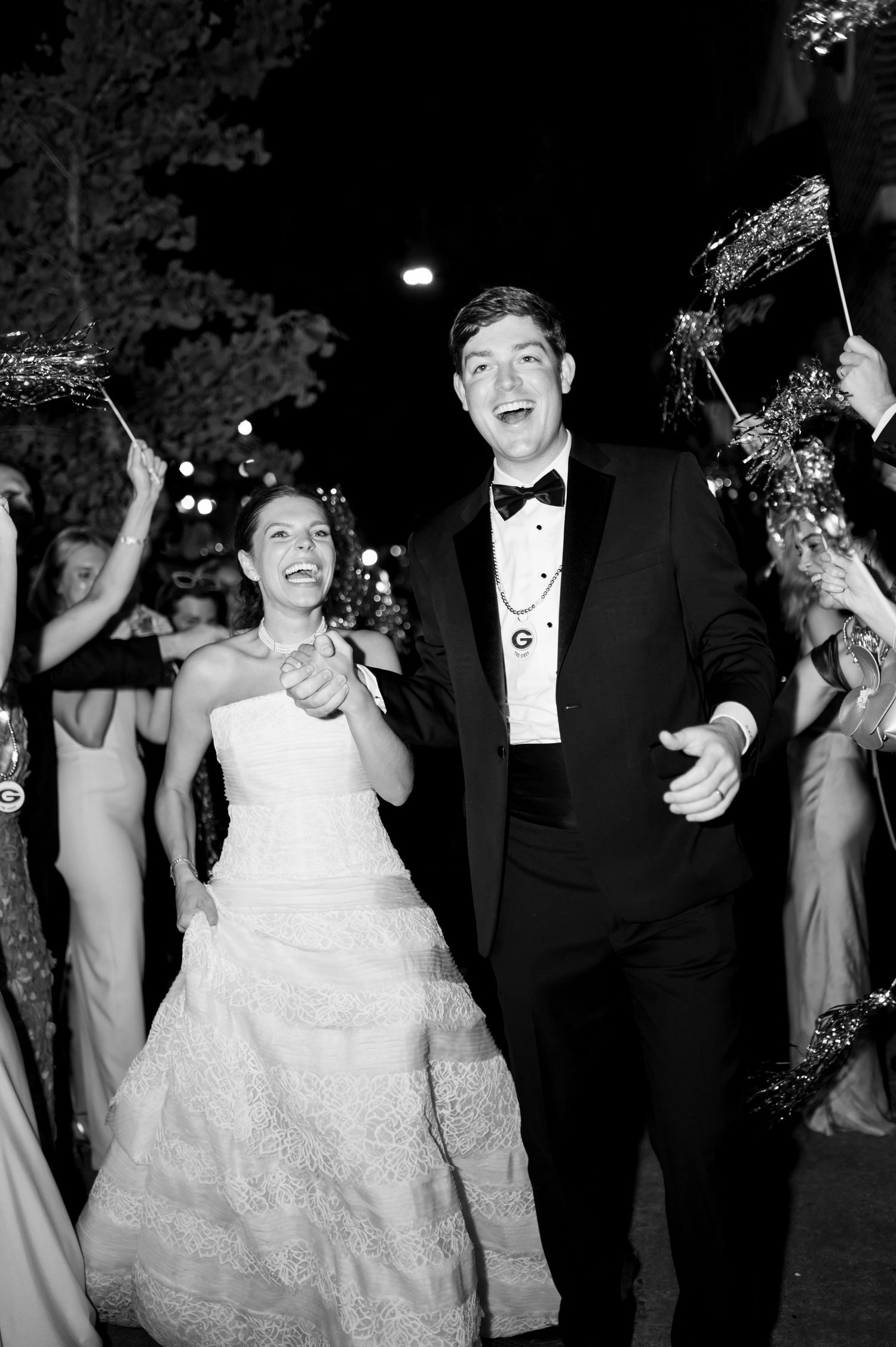 Black and white photo of joyful newlyweds exiting reception amid cheering guests with sparklers.