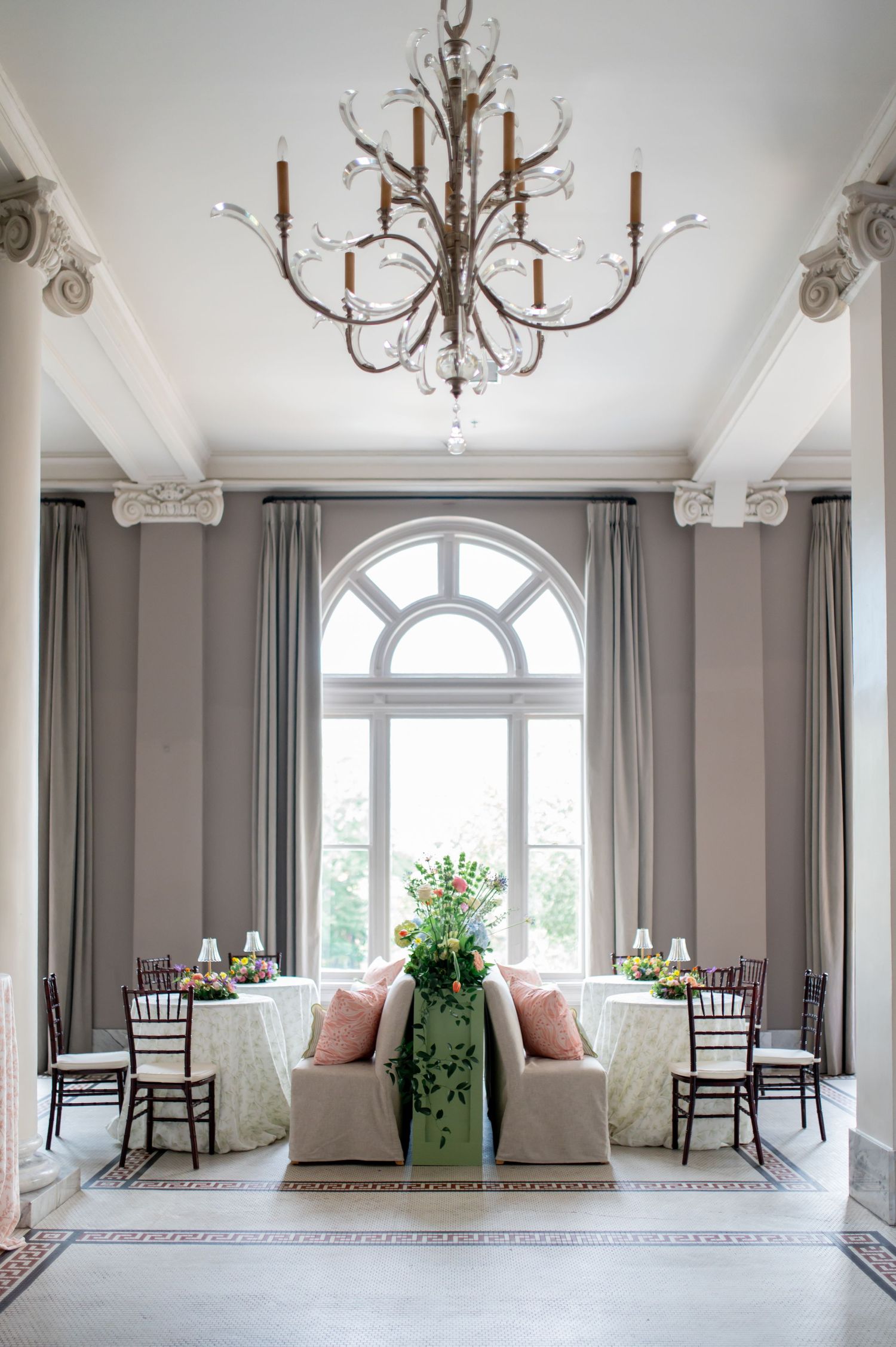 Elegant wedding reception room with chandelier, arched window, floral centerpiece, and chiavari chairs.