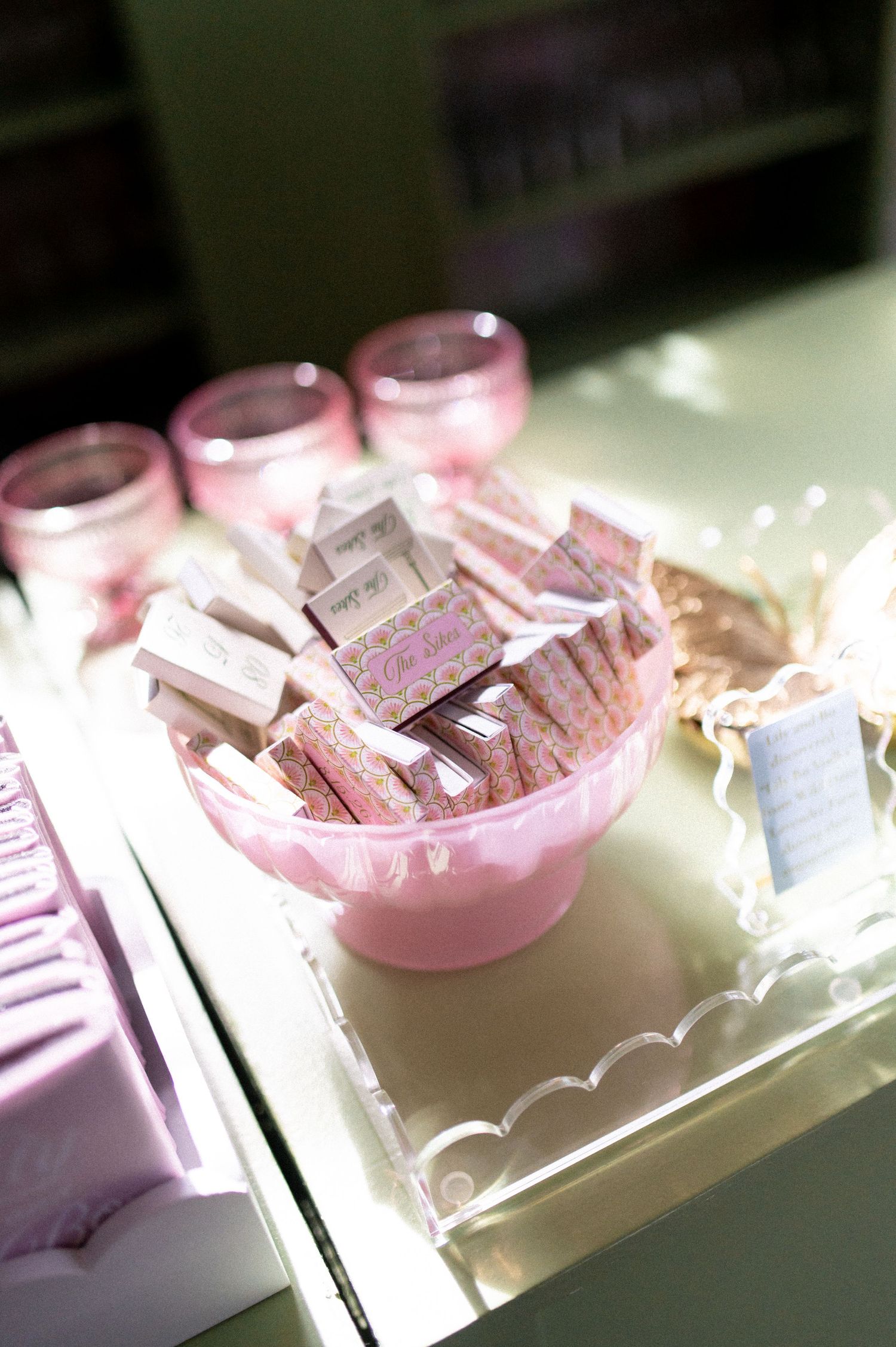 Pink cake stand filled with decorative wedding favor matchboxes on a scalloped acrylic tray.