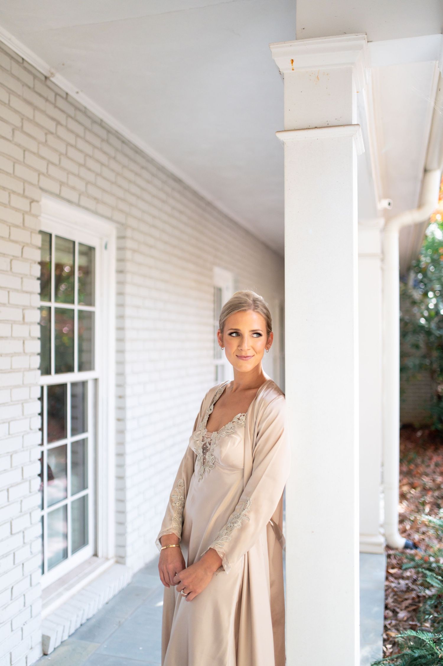Elegant figure in champagne silk robe standing on white columned porch.
