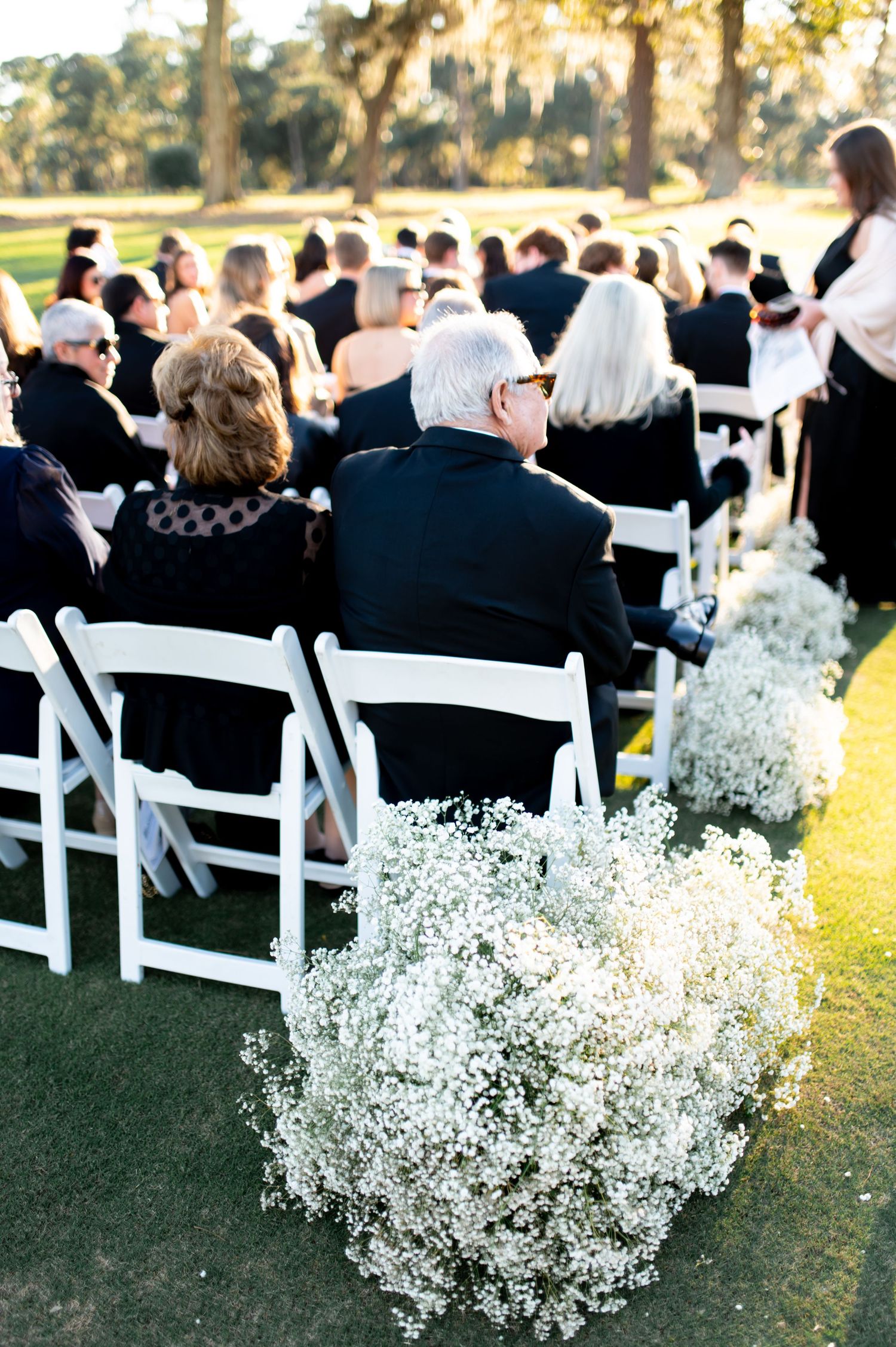 Wedding guests seated in white chairs with baby's breath floral arrangements lining the outdoor ceremony aisle.