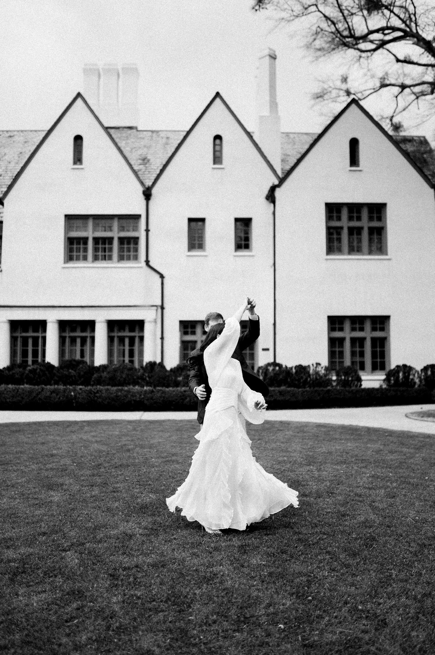 A bride in a flowing white wedding dress twirls gracefully on a lawn in front of white Tudor-style buildings.