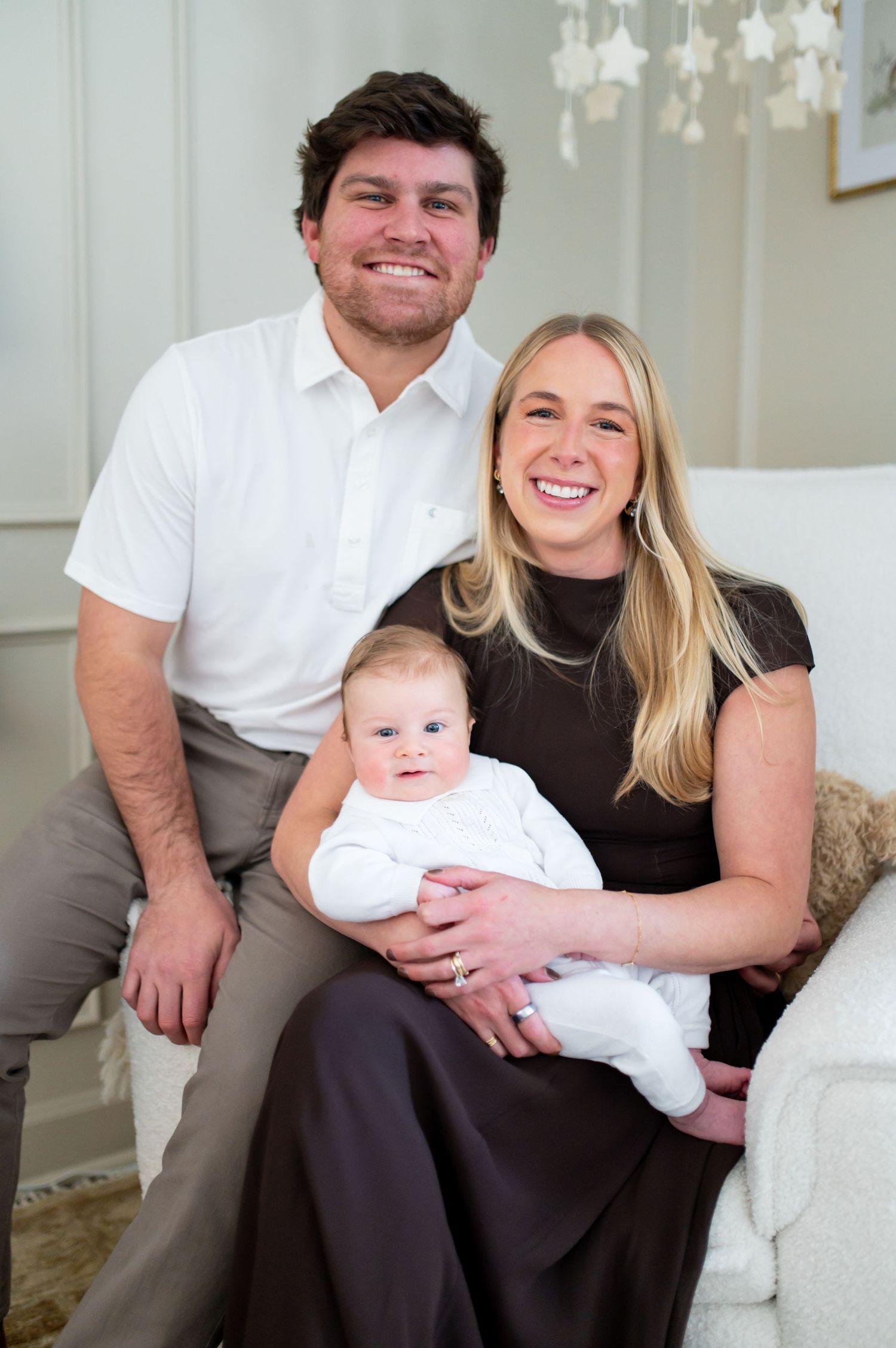 Family portrait series showing couple sitting together with baby in white outfit on light colored couch.