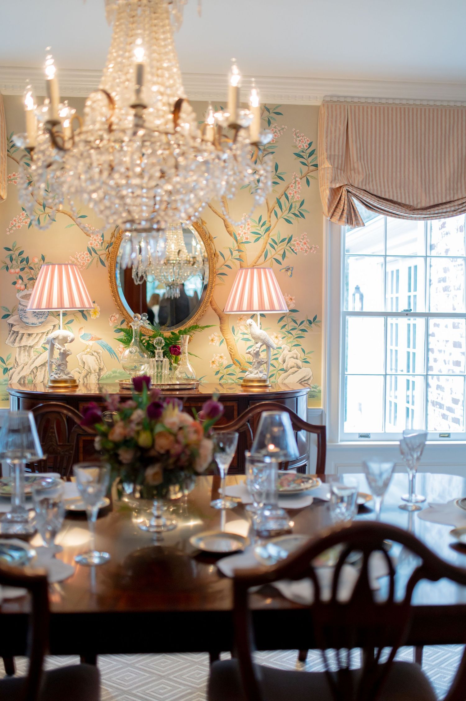 Traditional dining room with crystal chandelier, hand-painted floral wallpaper, buffet with pink lamps, and set table.