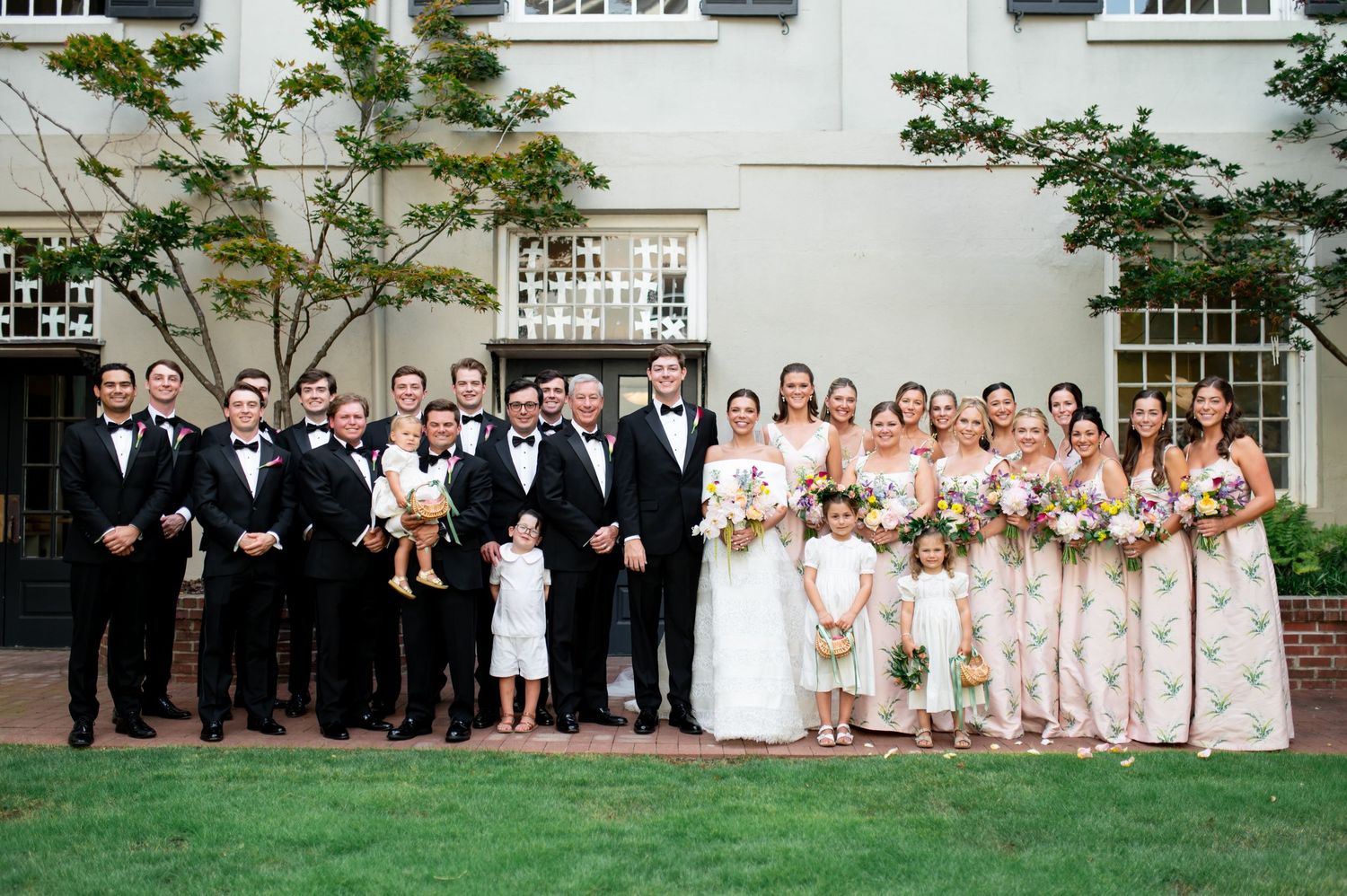 Large wedding party posing outdoors in formal attire with bridesmaids in floral dresses.