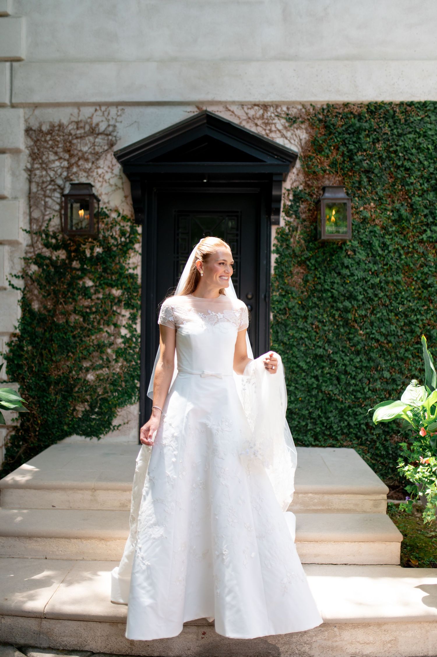 Elegant bride in lace cap sleeve wedding gown stands on steps surrounded by ivy-covered walls.