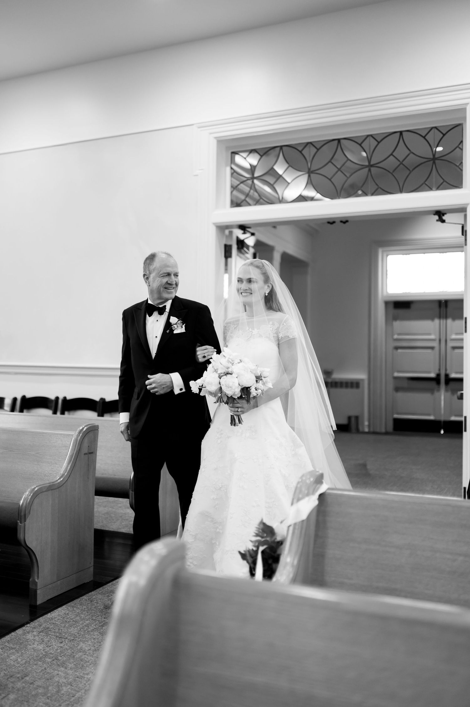 Father escorts bride down church aisle in classic black and white wedding photo.