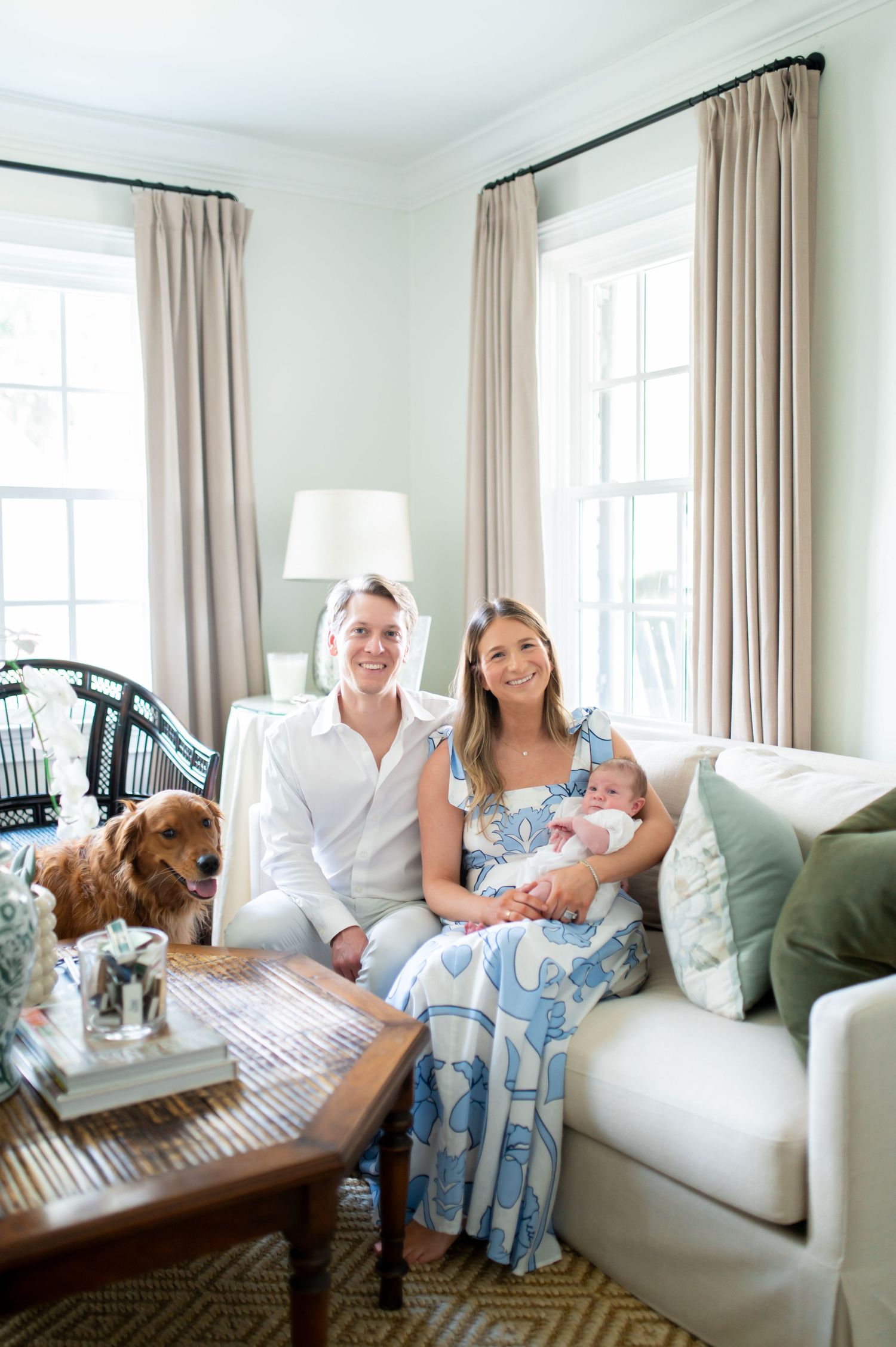 A family relaxes together on a white couch in a bright, airy living room with beige curtains and natural lighting.