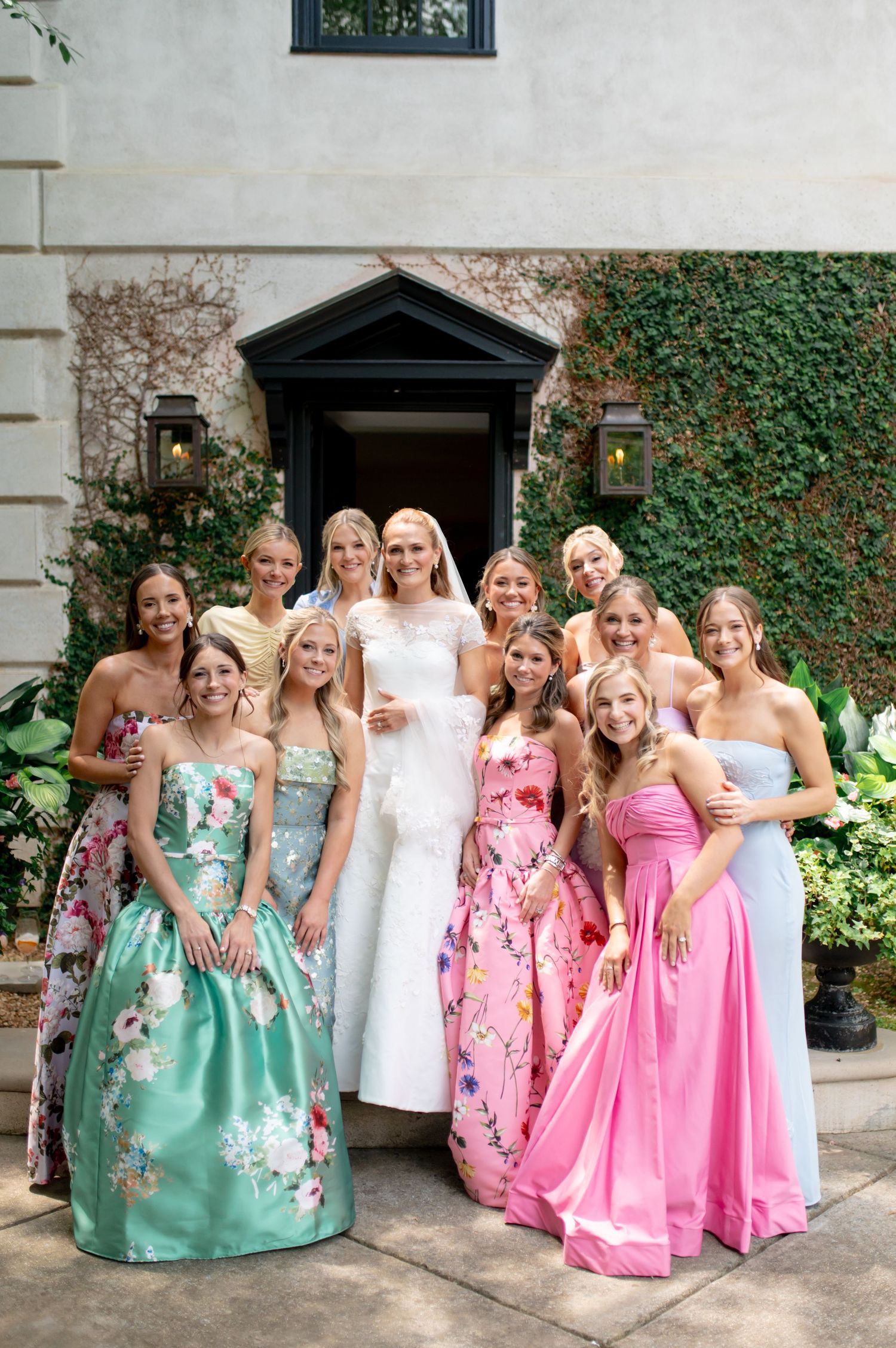 Bride in white wedding gown poses with bridesmaids in colorful floral dresses outside elegant venue.