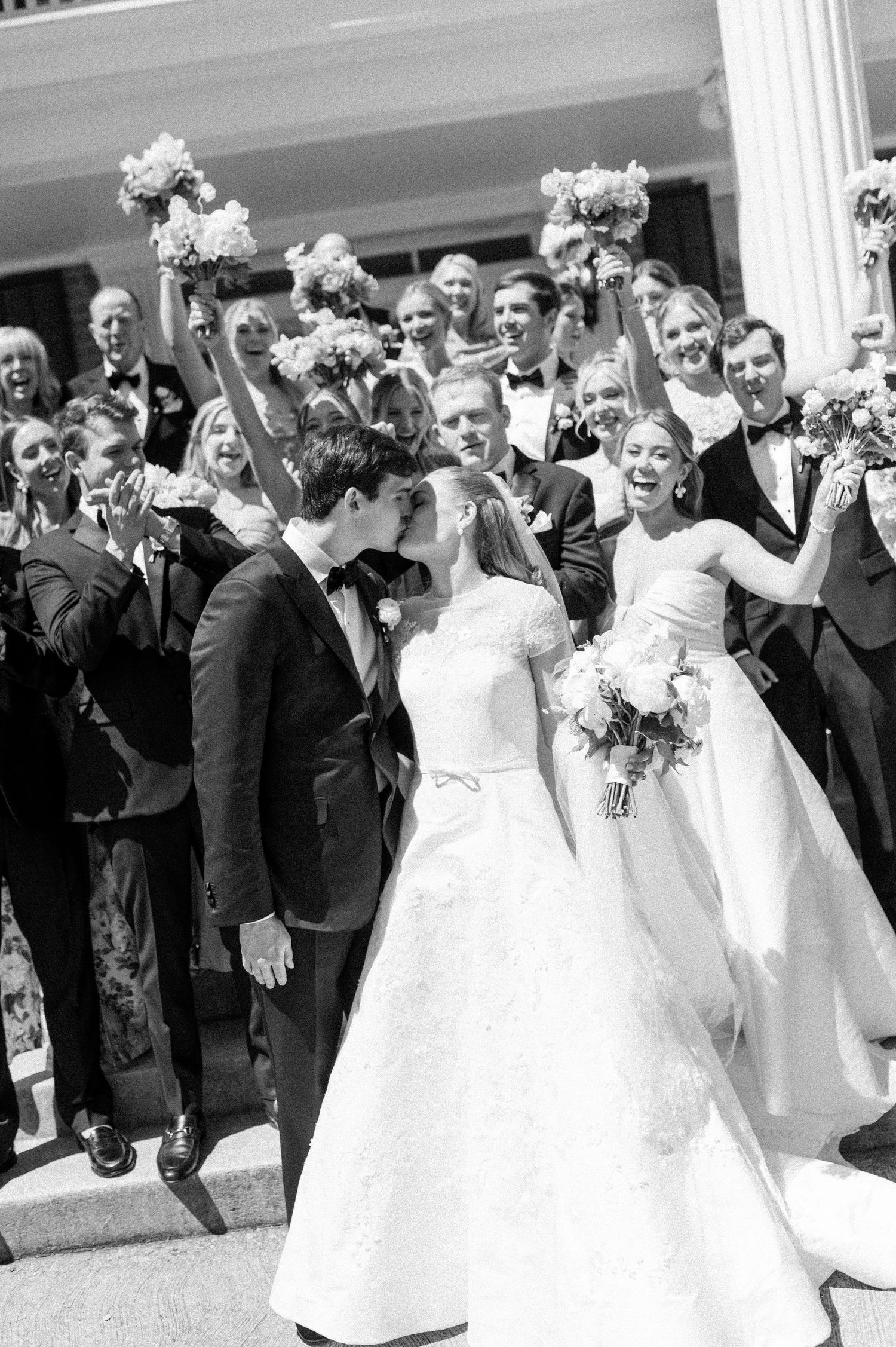 Black and white photo of newlyweds kissing as wedding party celebrates with raised bouquets behind them.