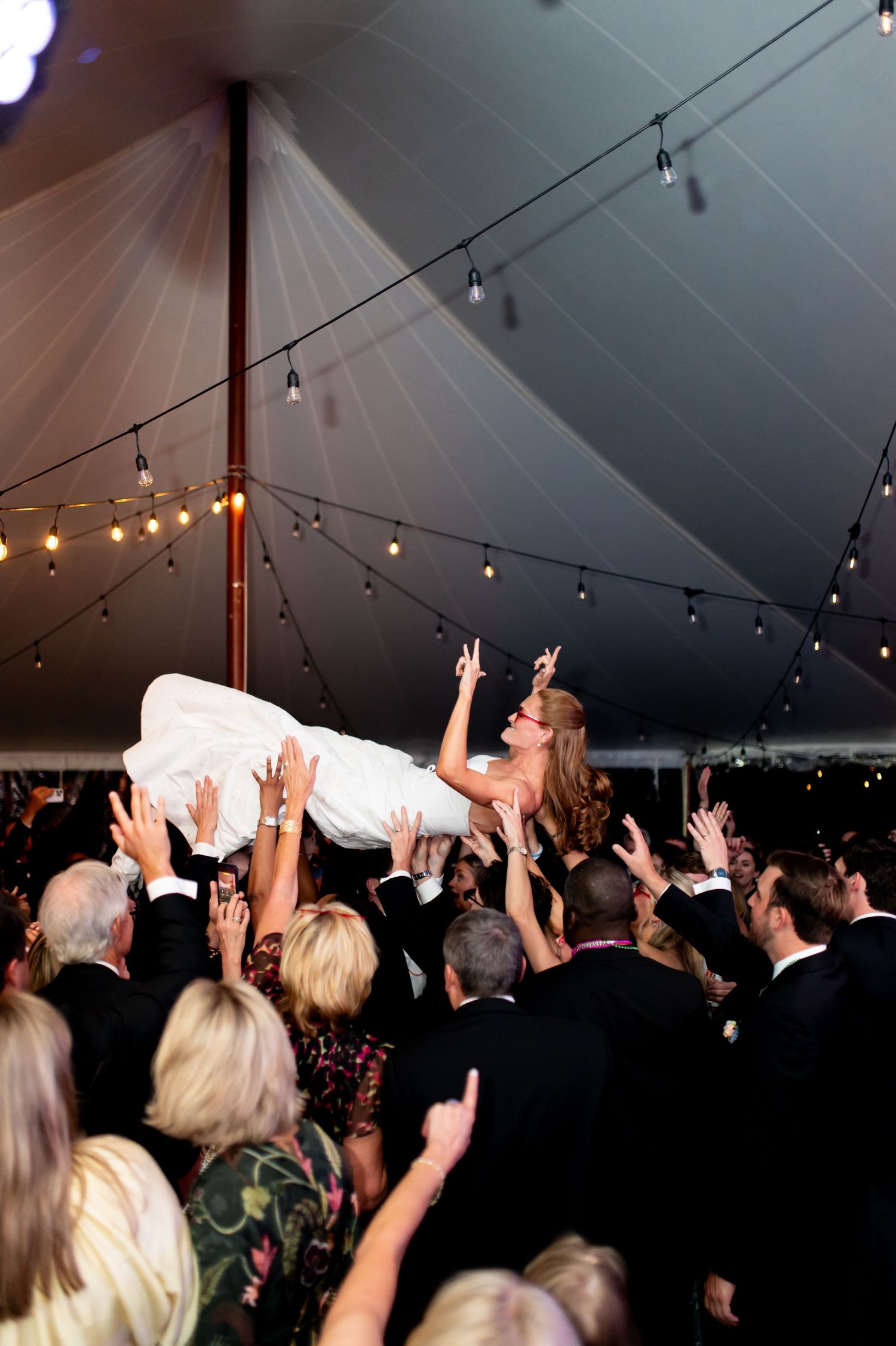 Bride crowd surfs above cheering wedding guests under string lights inside elegant white reception tent.