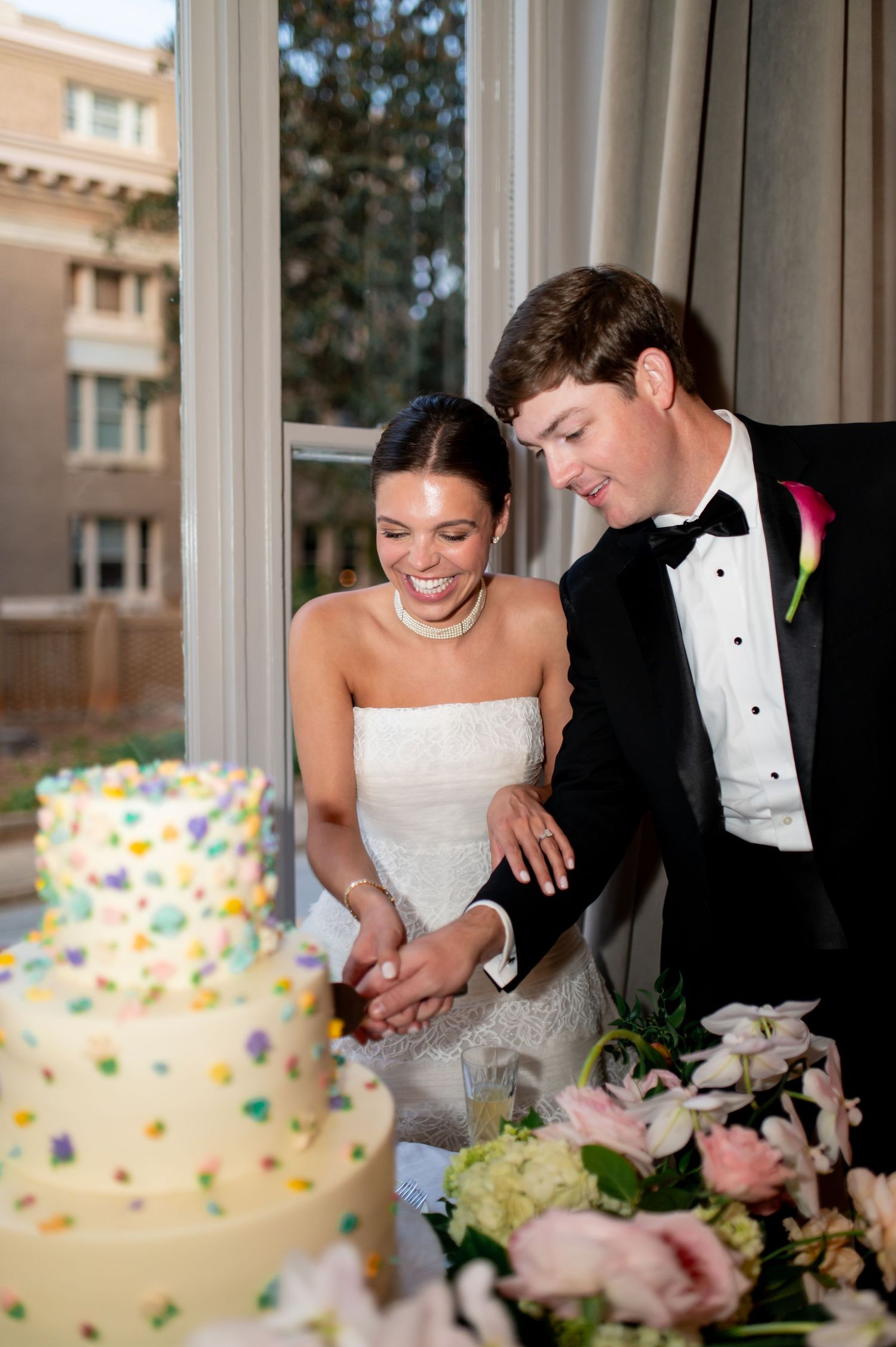 Bride and groom smile while cutting colorful confetti-dotted tiered wedding cake at reception.