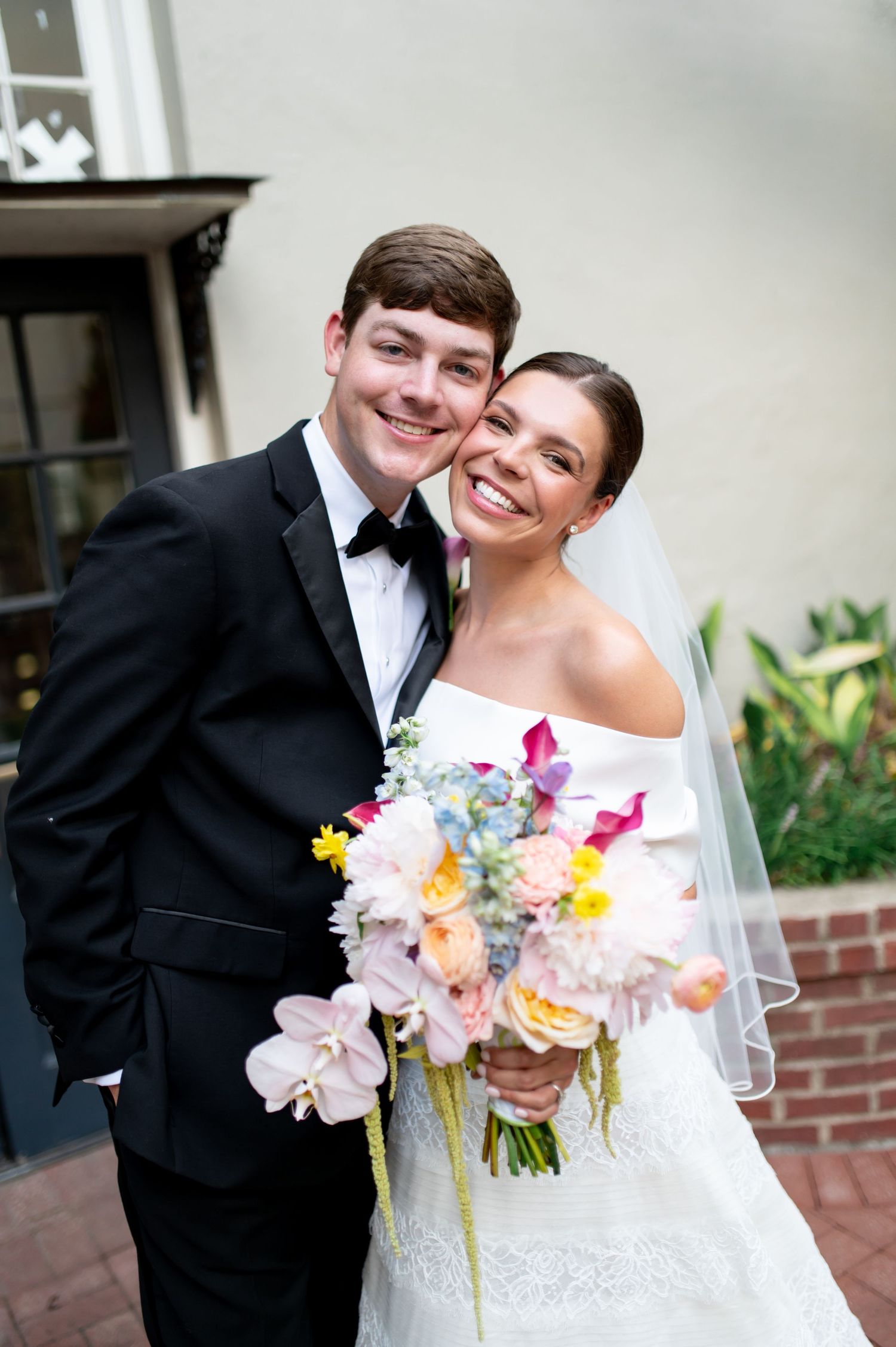 Smiling bride and groom posing together outdoors, bride holding a colorful spring wedding bouquet.