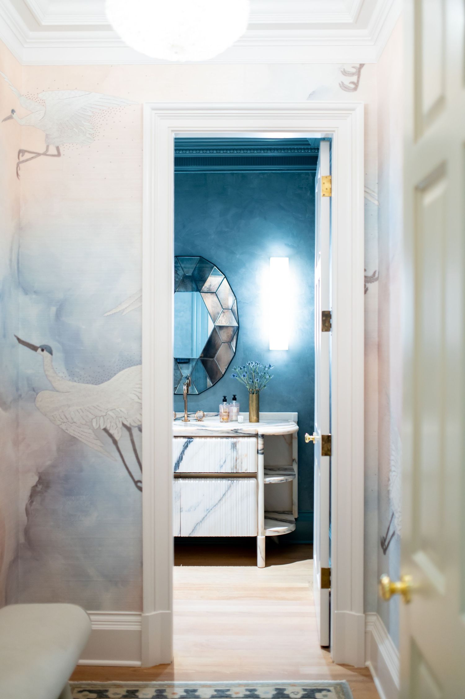 Elegant hallway with crane mural wallpaper leading to a blue bathroom with marble vanity and geometric mirror.
