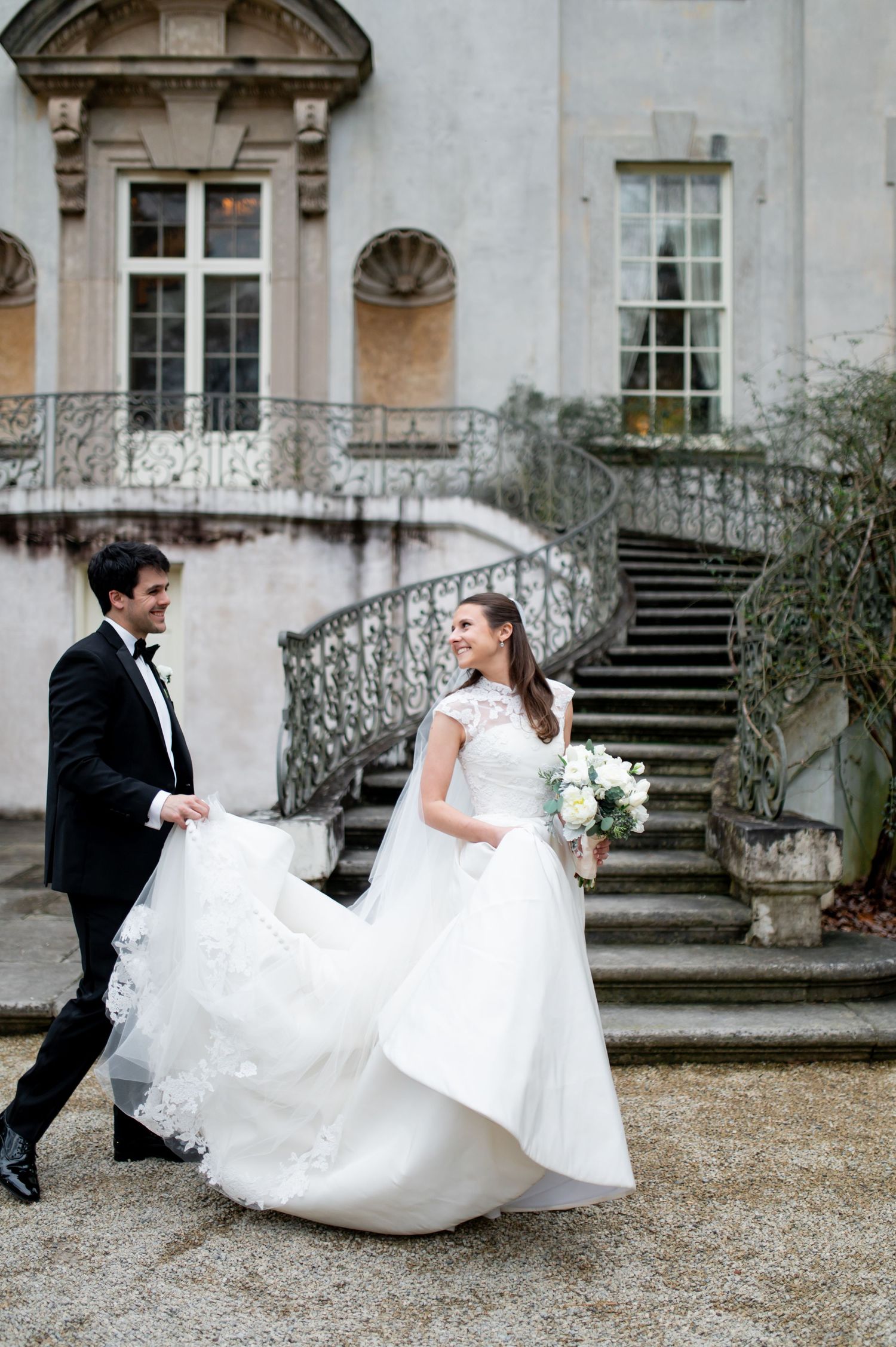 A bride and groom walk joyfully outside a grand estate, her white lace gown flowing as he holds her train.