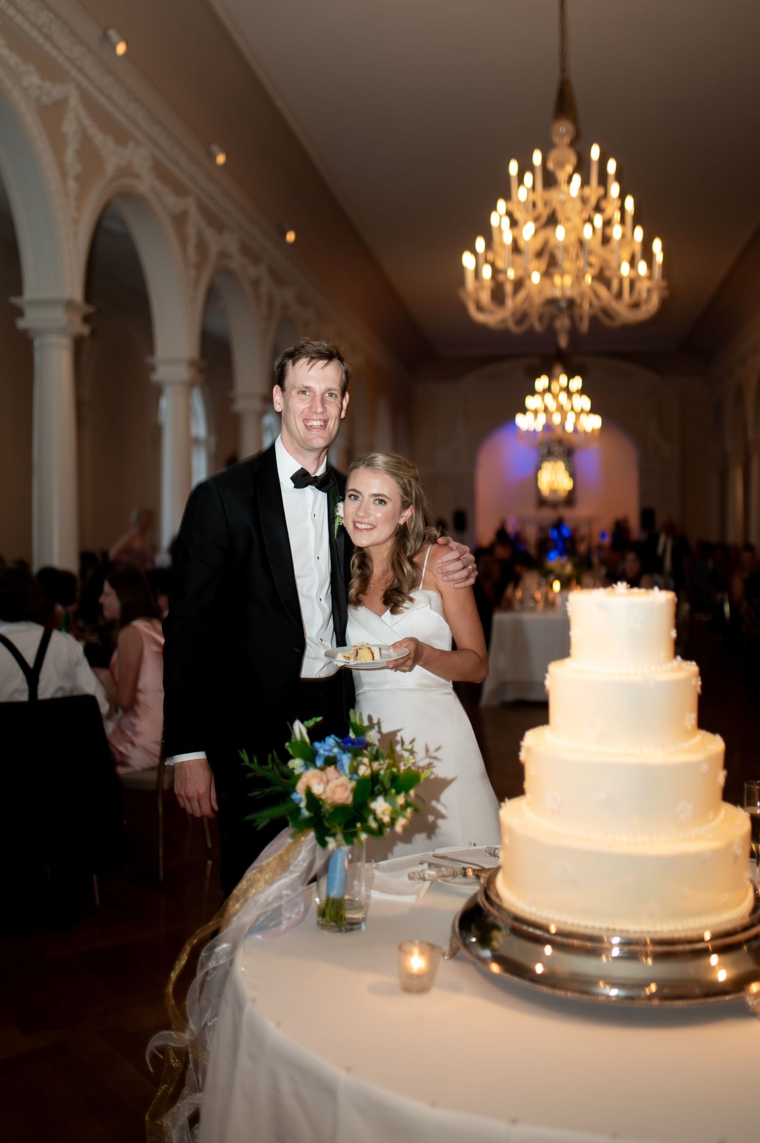 Elegant white tiered wedding cake displayed in a grand ballroom with chandeliers.