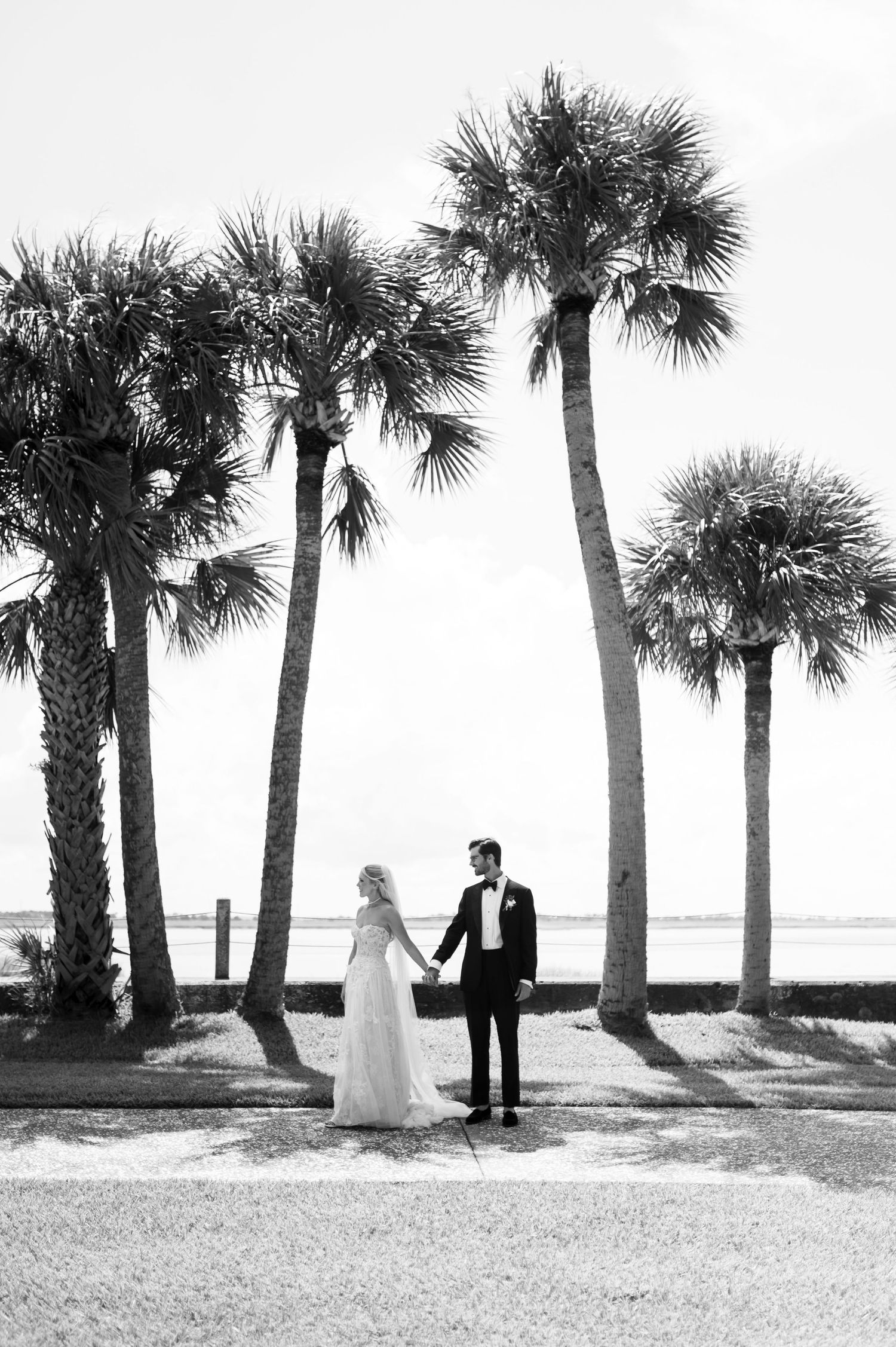Wedding couple walks among palm trees on beach in black and white photograph.