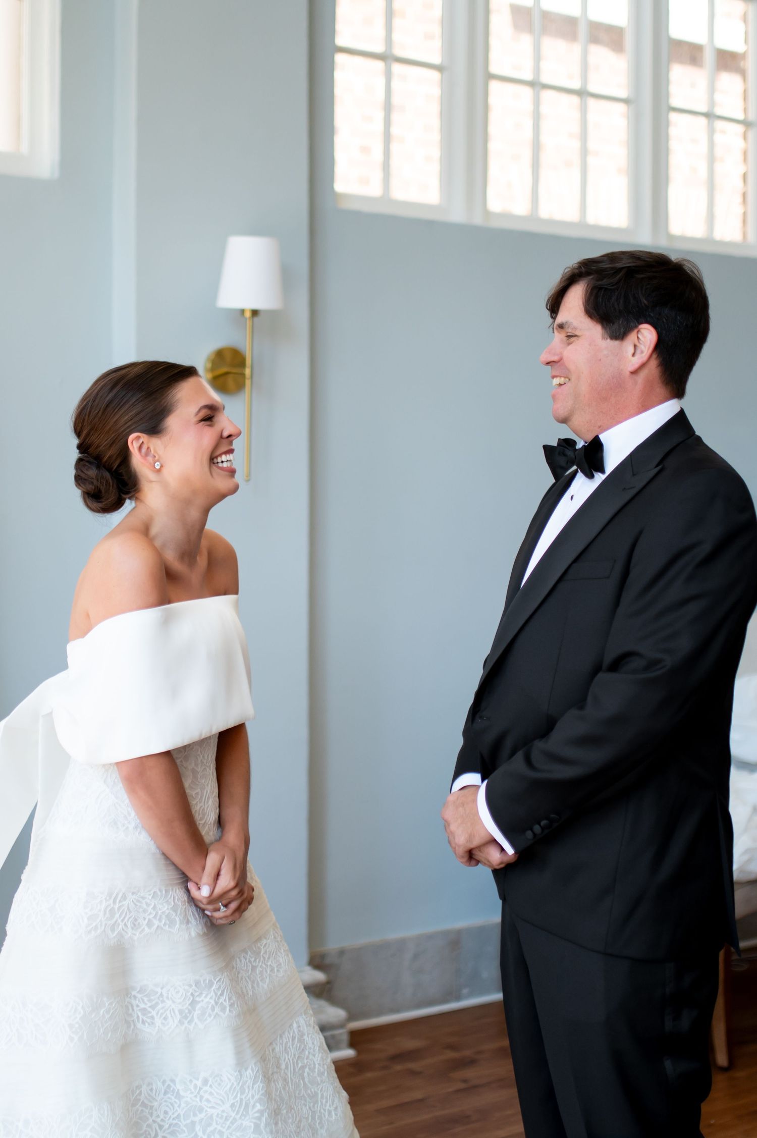 Bride in off-shoulder white gown laughs with man in black tuxedo during elegant wedding first look.