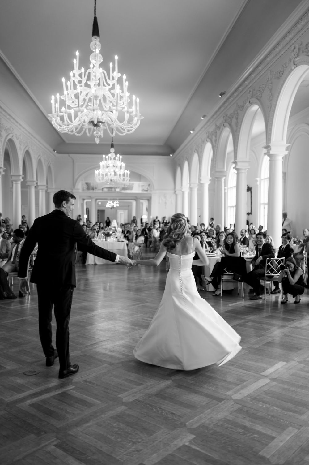 A couple dances in an elegant ballroom with crystal chandeliers and arched columns during their wedding reception.