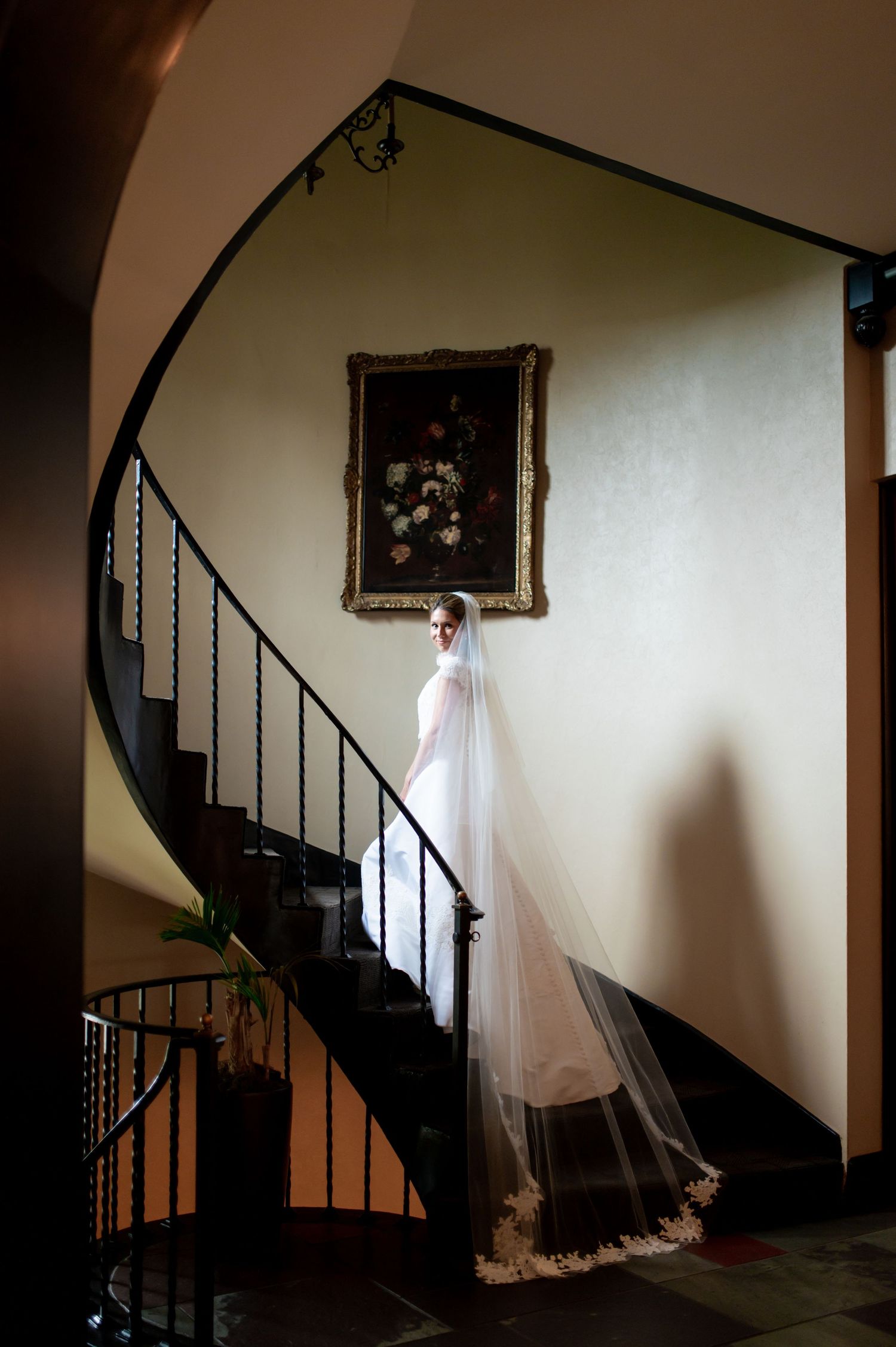 Wedding dress photographed near spiral staircase with dramatic lighting and framed artwork.
