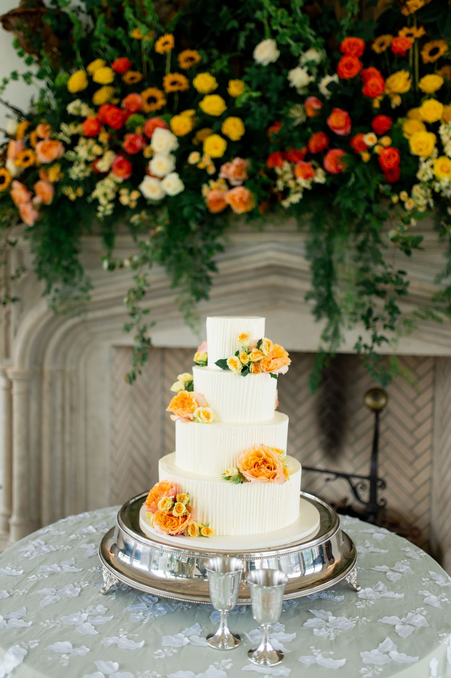 White wedding cake adorned with orange flowers displayed on silver stand beneath floral-decorated archway.