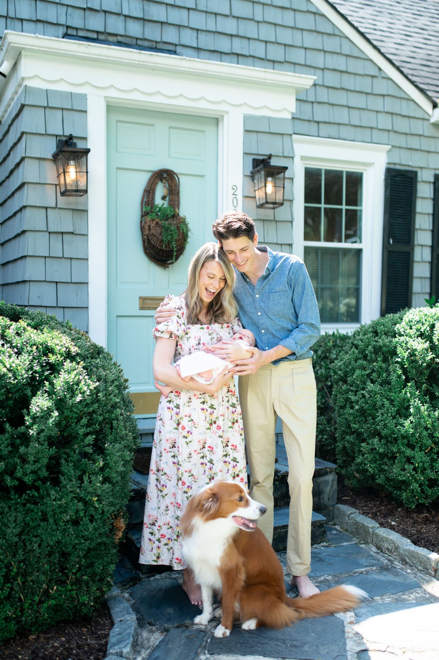 A couple stands with their newborn baby outside a gray house with a blue door and a brown dog sitting beside them.