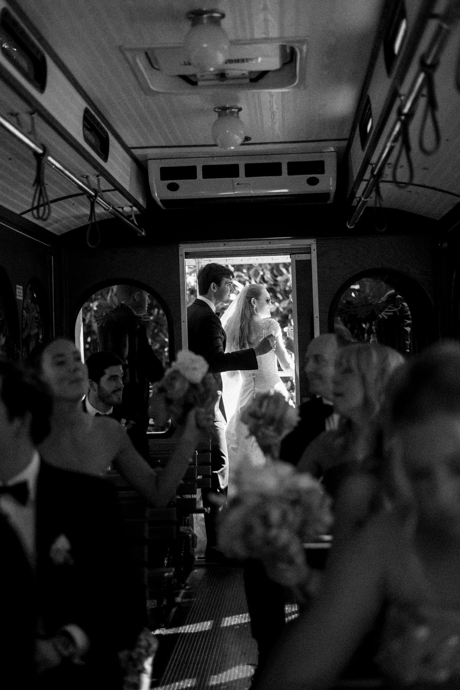 Wedding guests celebrate as bride and groom stand at bus entrance in black and white photo.