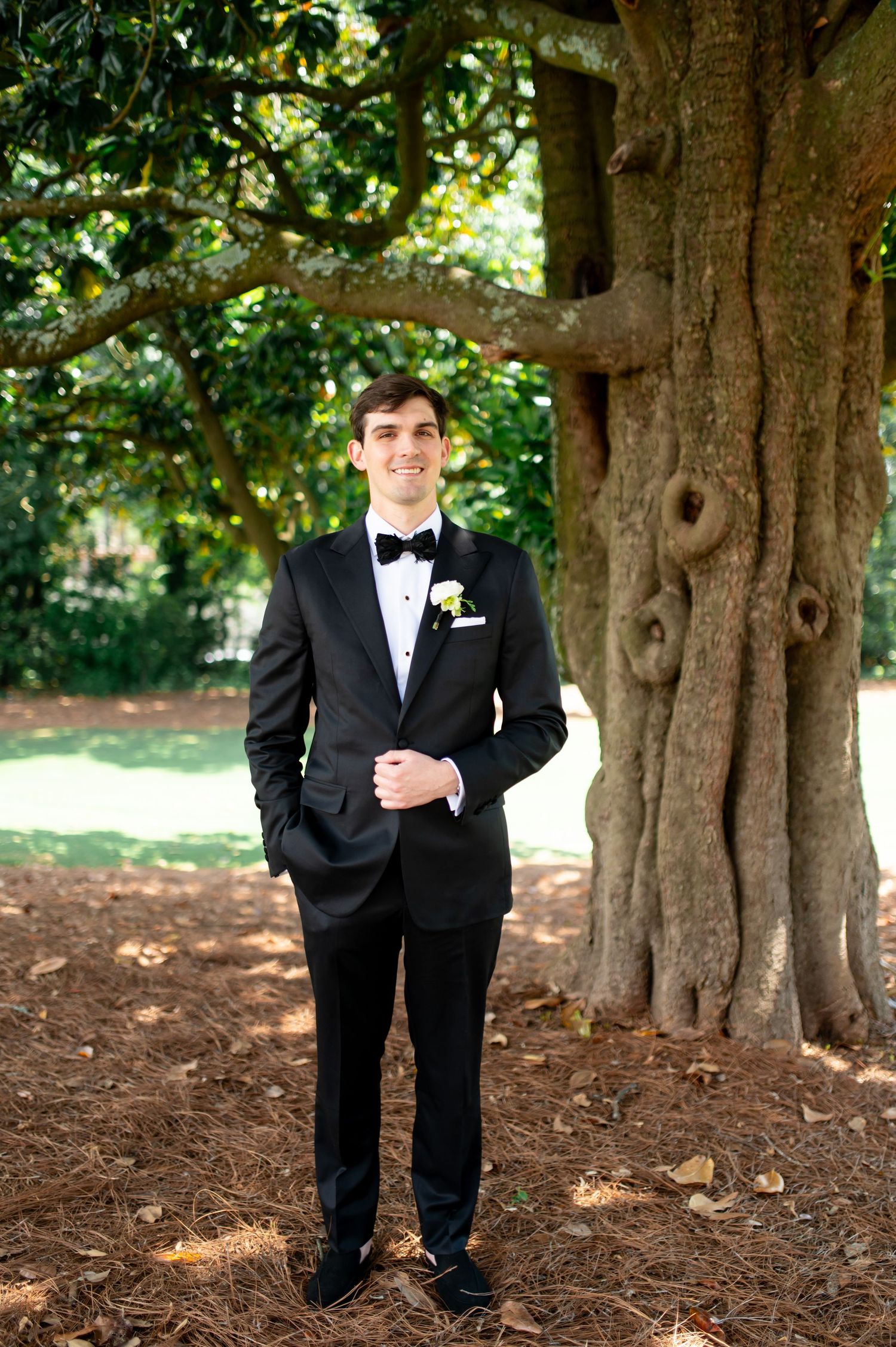 Dapper groom in black tuxedo with boutonniere stands confidently beneath a large tree outdoors.