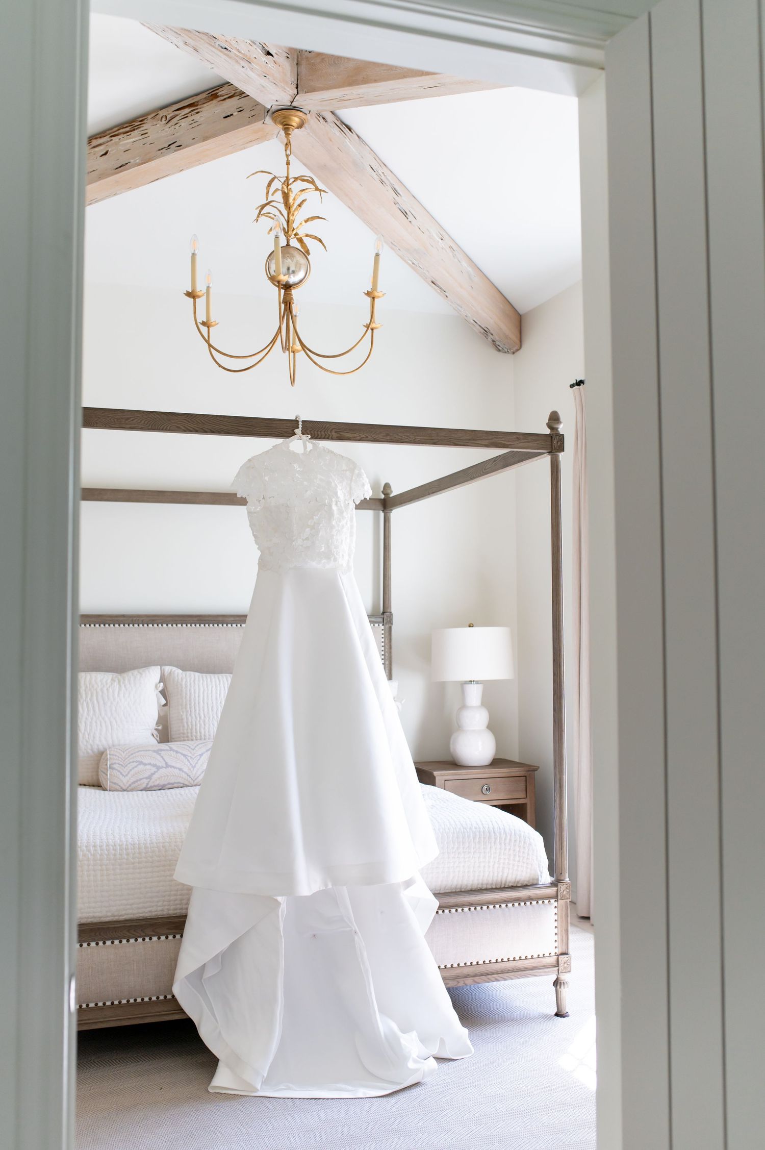 White wedding dress hanging from a gold chandelier in an elegant bedroom with wooden beams and a four-poster bed.