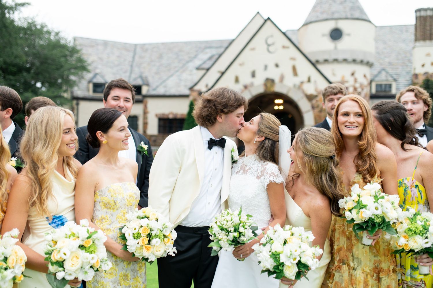 Wedding party wearing yellow and white dresses pose on a green lawn in front of a stone church.