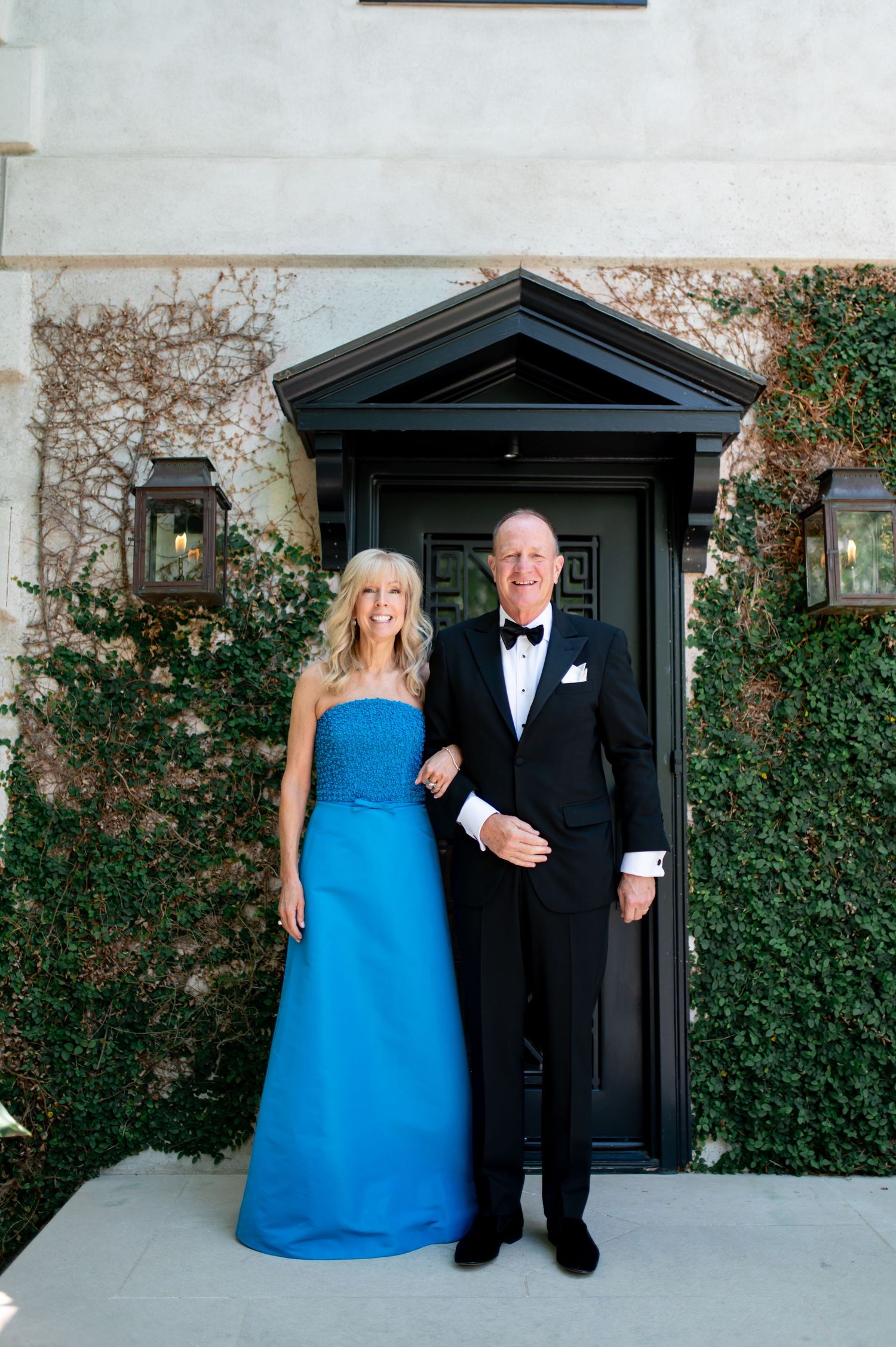 Elegant couple in formal attire, woman in blue strapless gown and man in tuxedo, pose at ivy-covered door.