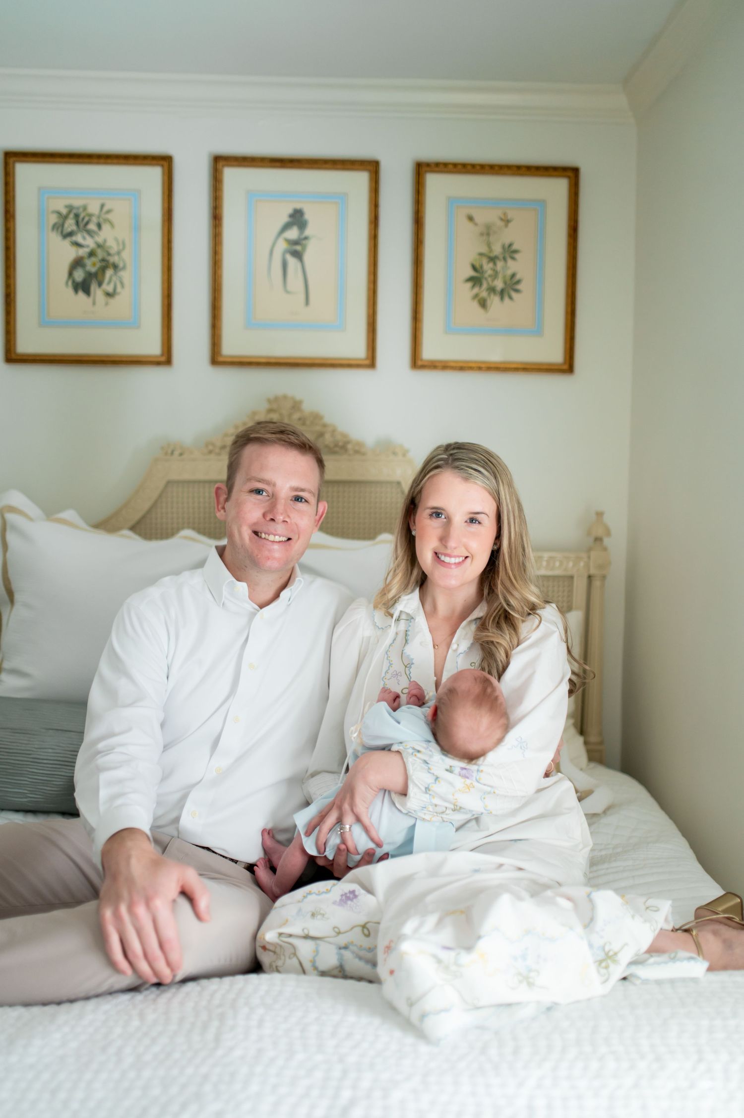 A couple in white clothing reclines together on a bed with framed artwork visible on the wall behind them.