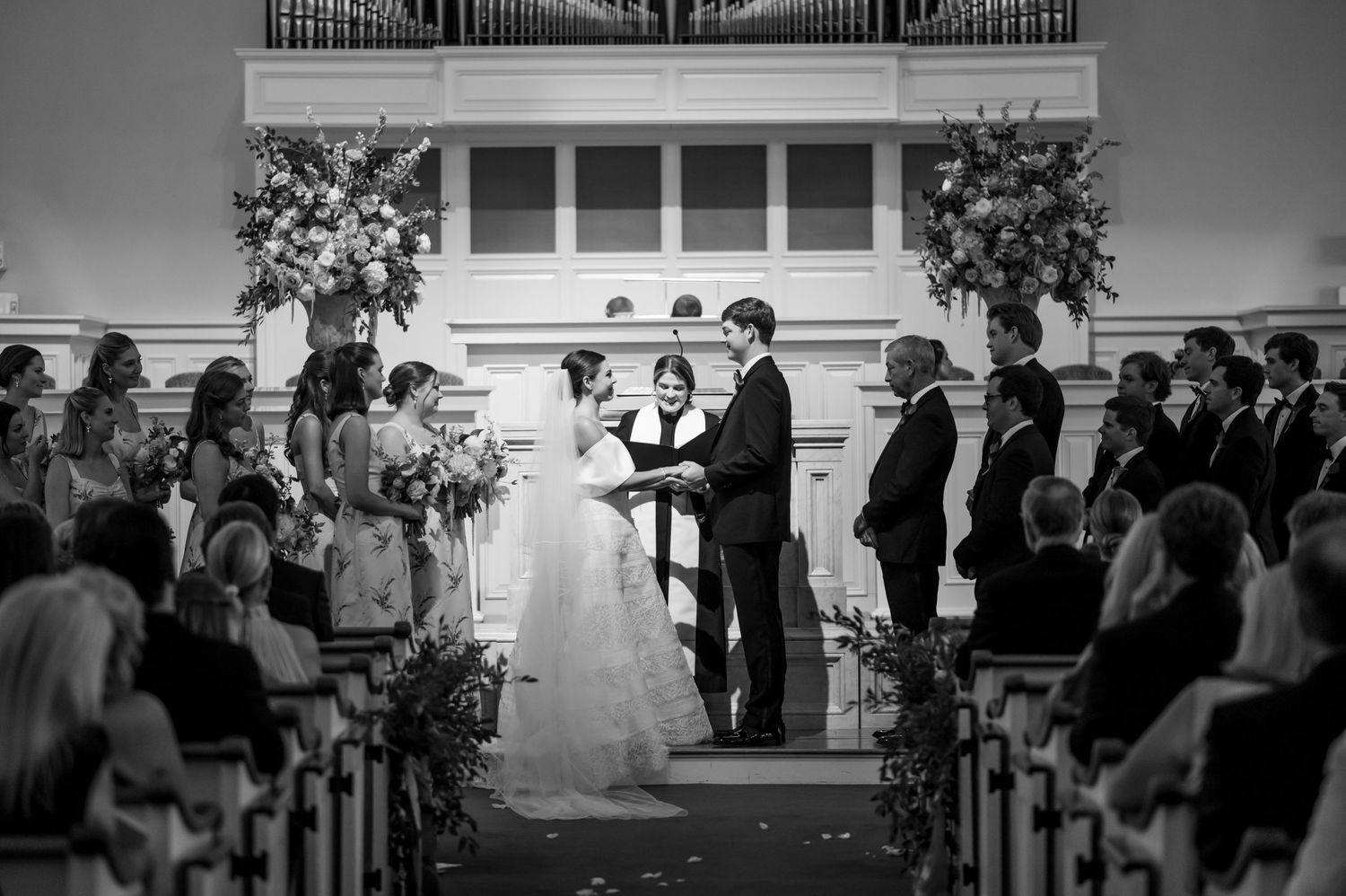 Black and white photo of a bride and groom exchanging vows at a church altar surrounded by wedding party.