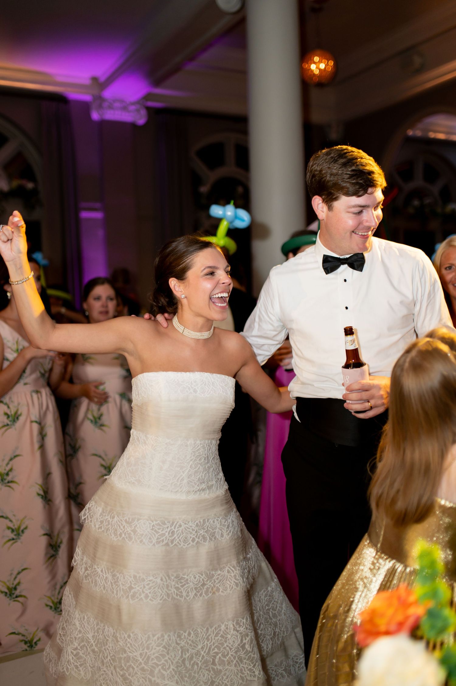 Laughing bride in lace gown dances with groom in bow tie on crowded wedding reception floor.