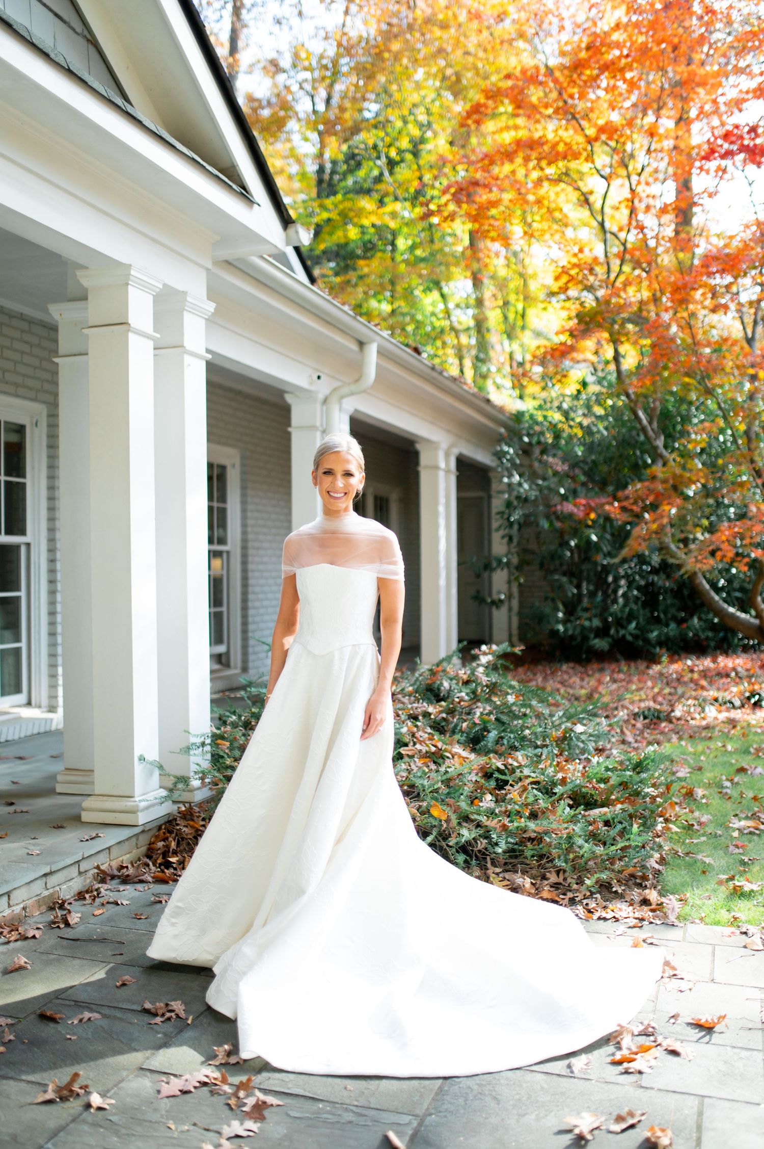 A stunning white wedding gown with a long train photographed on a porch with autumn foliage in the background.